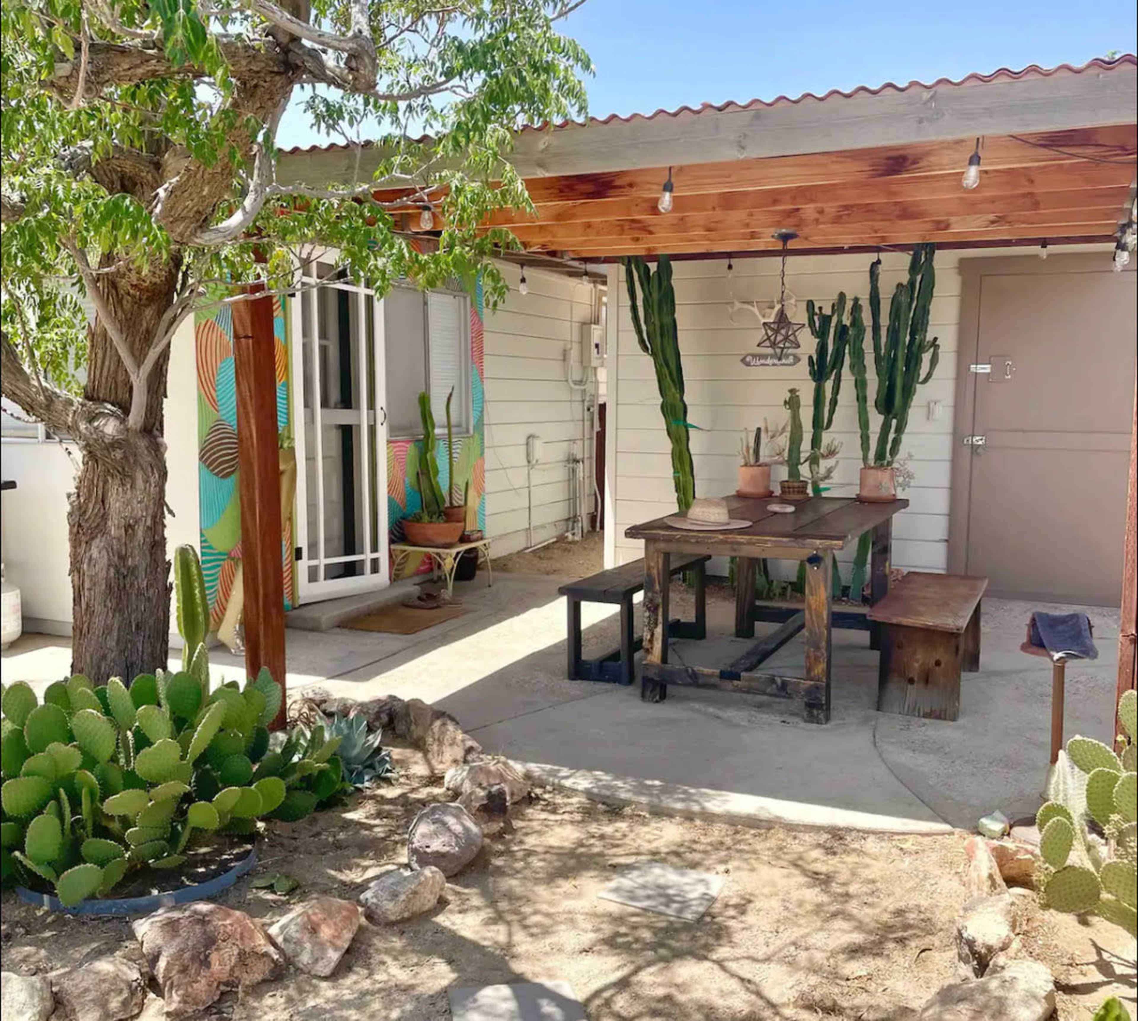 The image shows a patio area with a wooden table and benches under a roof, surrounded by potted cacti and a garden featuring stones and plants.