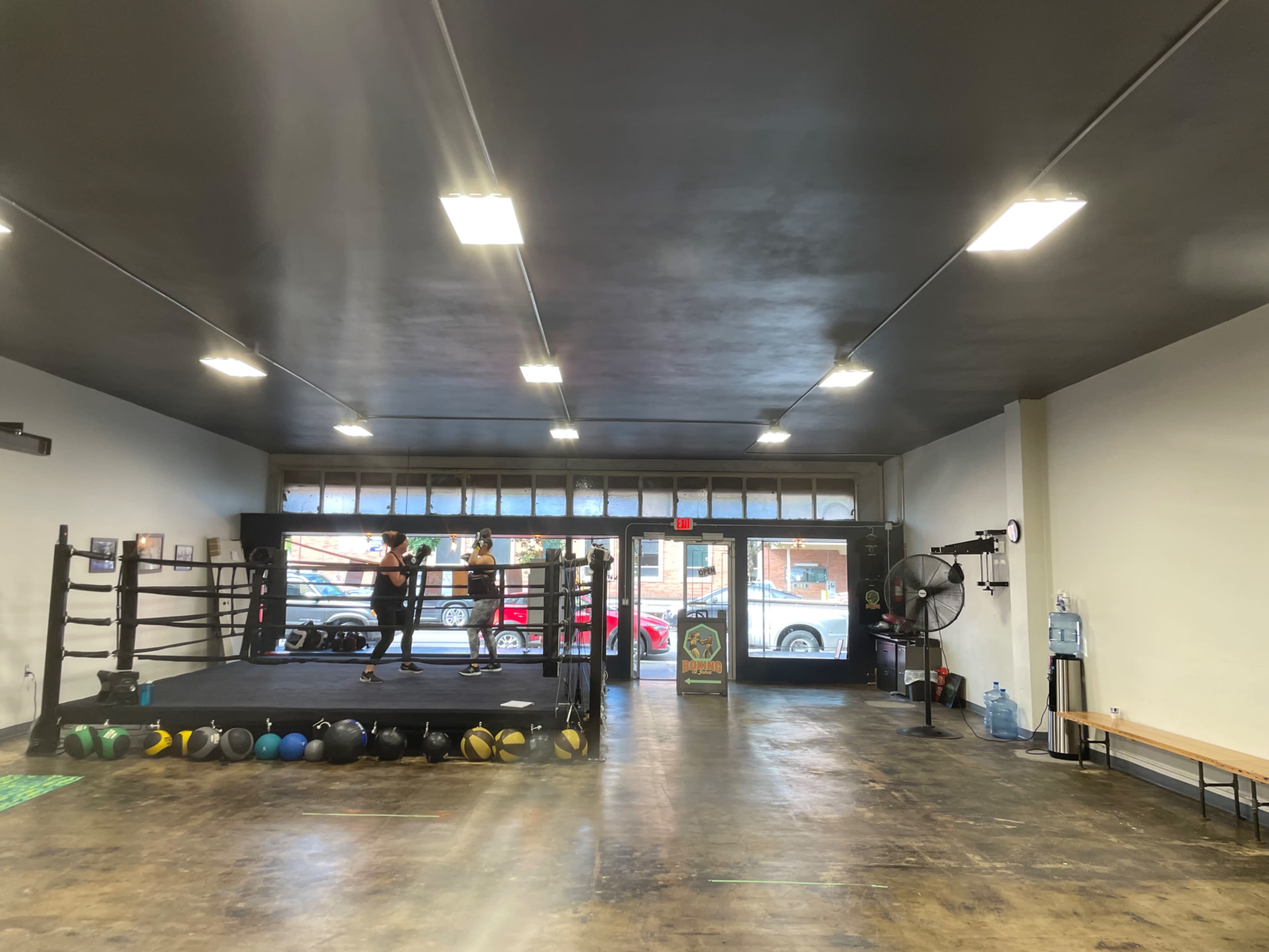 An indoor boxing gym with a boxing ring, training equipment, and large windows facing the street.