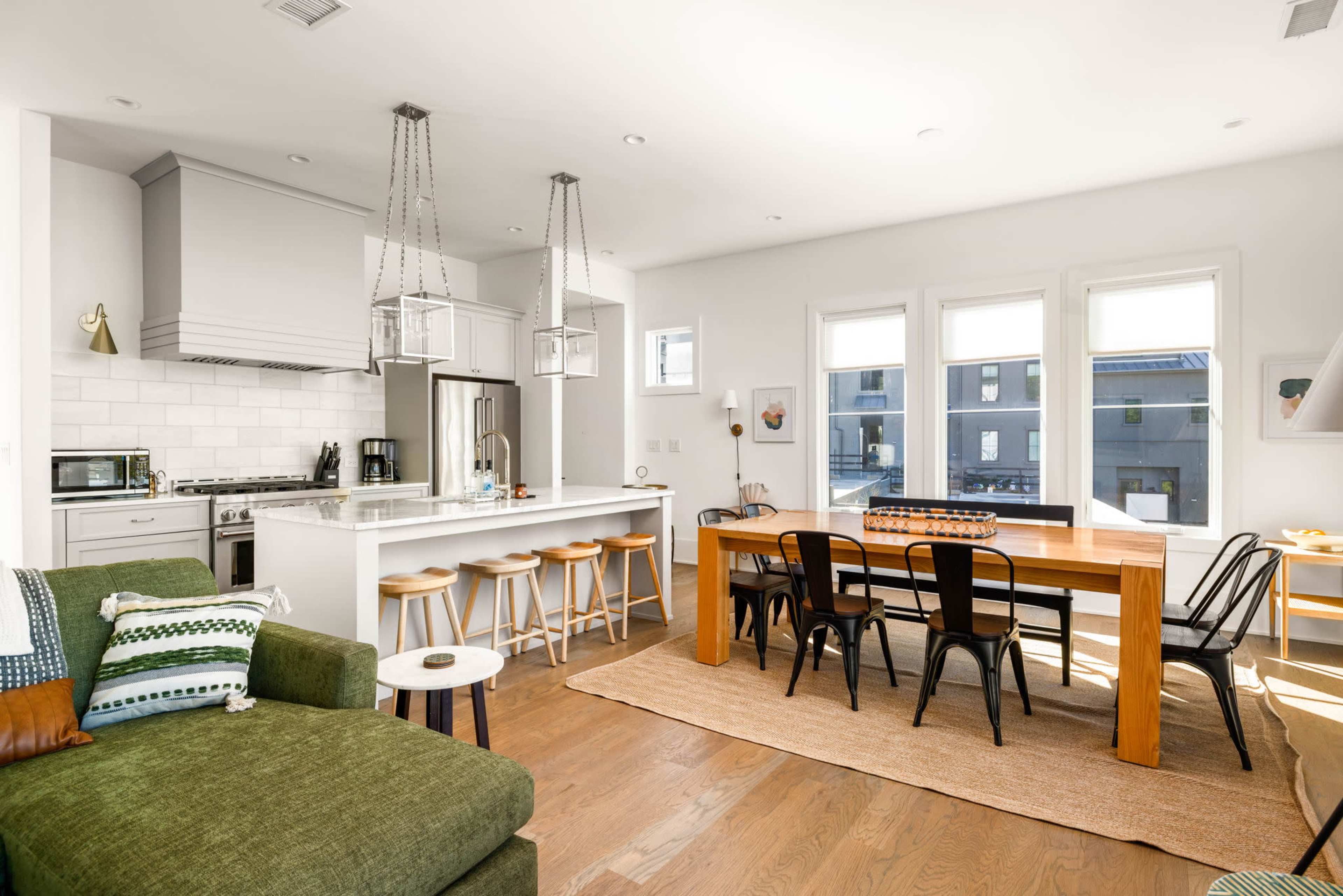 The image shows a modern open-concept kitchen and dining area featuring a large wooden table, bar stools, and a green couch in the living space.