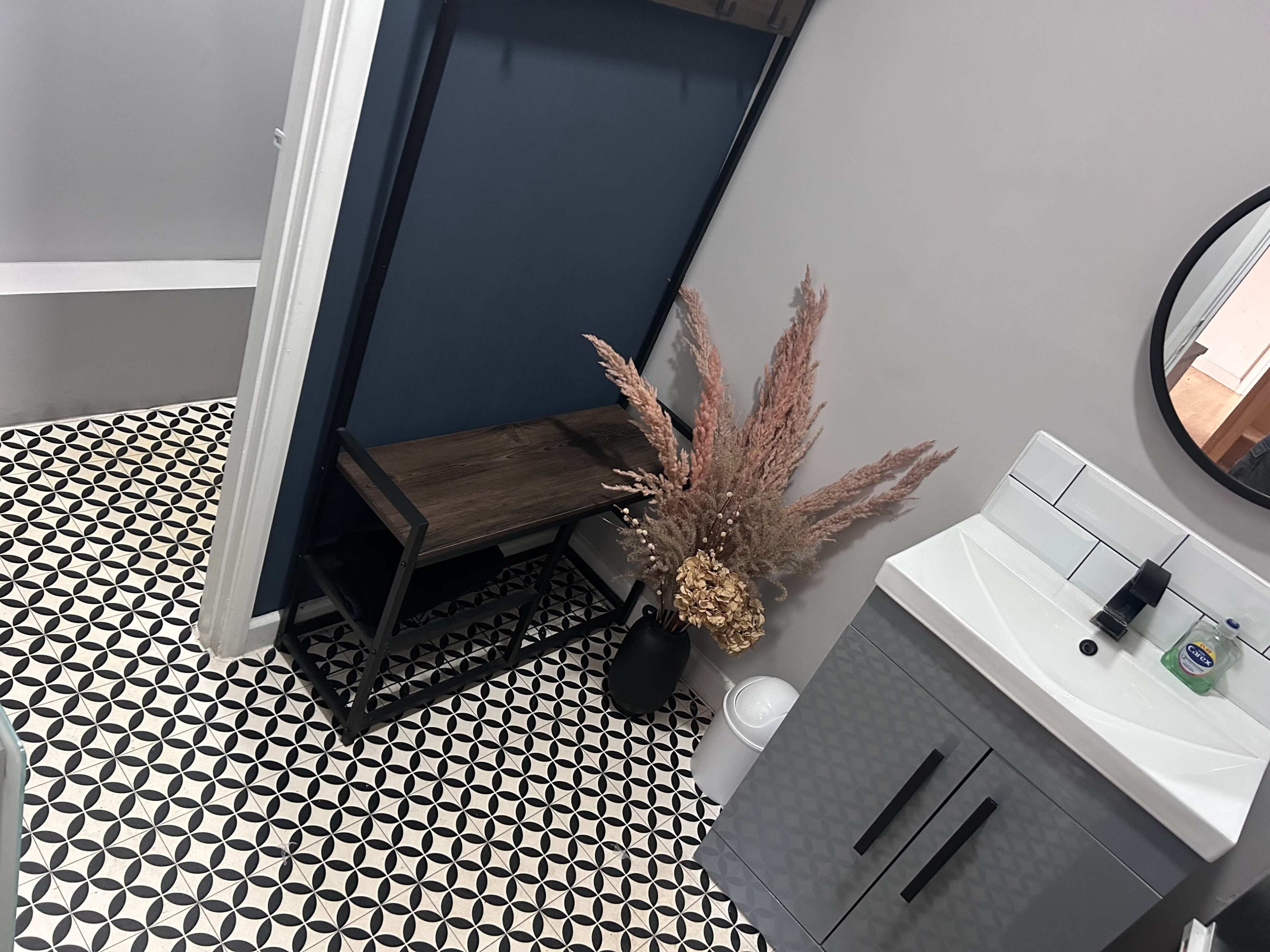 A bathroom featuring a patterned floor, a wooden bench against a dark blue wall, a round mirror, and a sink with a cabinet below.