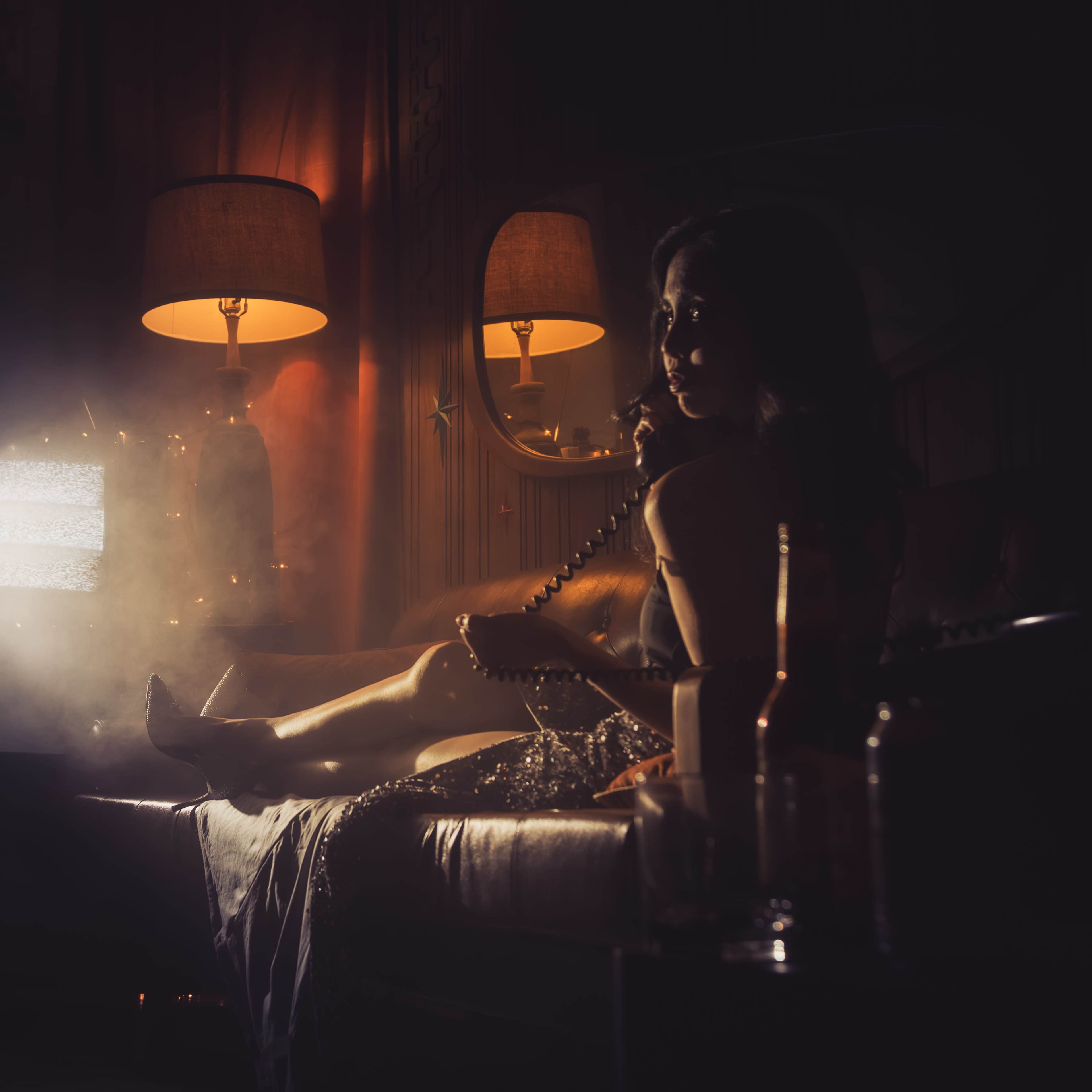 A woman sits on a sofa in a dimly lit room, talking on a vintage phone while illuminated by two table lamps and a flickering television.