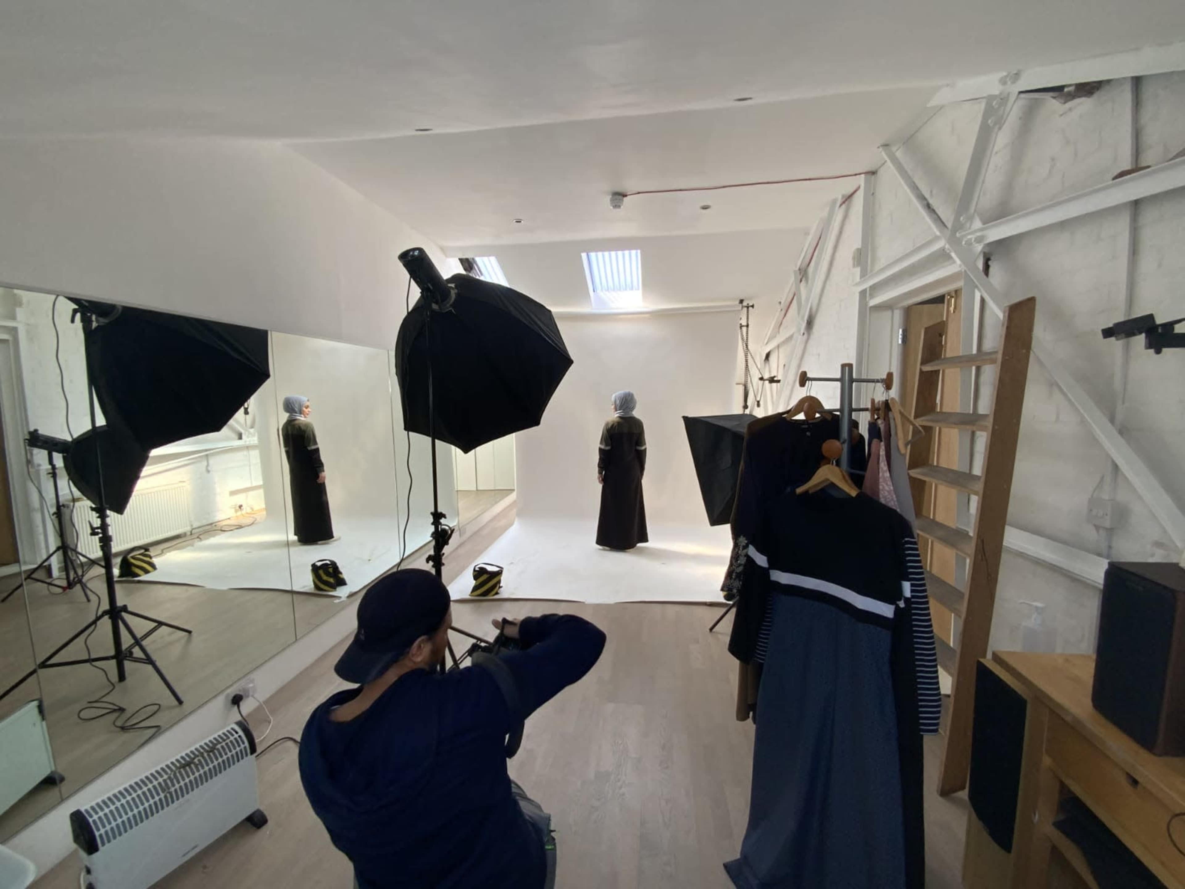 A photographer captures a model in a studio with a large mirror and clothing hanging on a rack in the background.