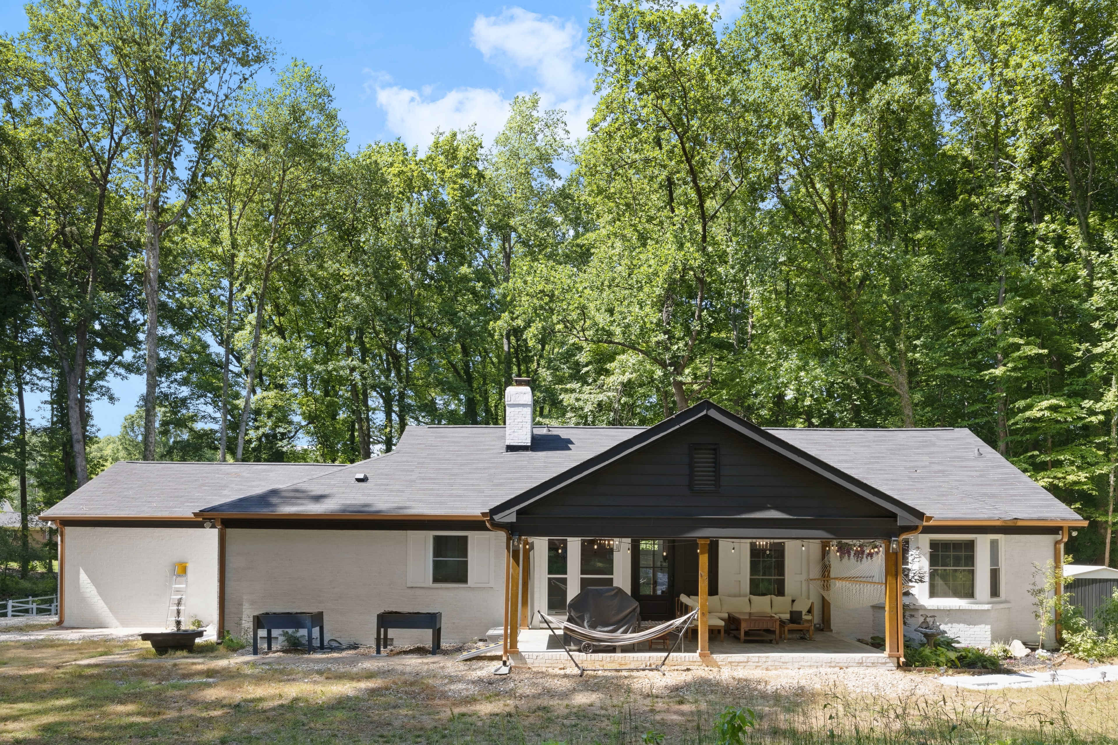 The image shows a single-story house surrounded by tall trees, featuring a combination of white and dark exterior accents along with a covered outdoor seating area.