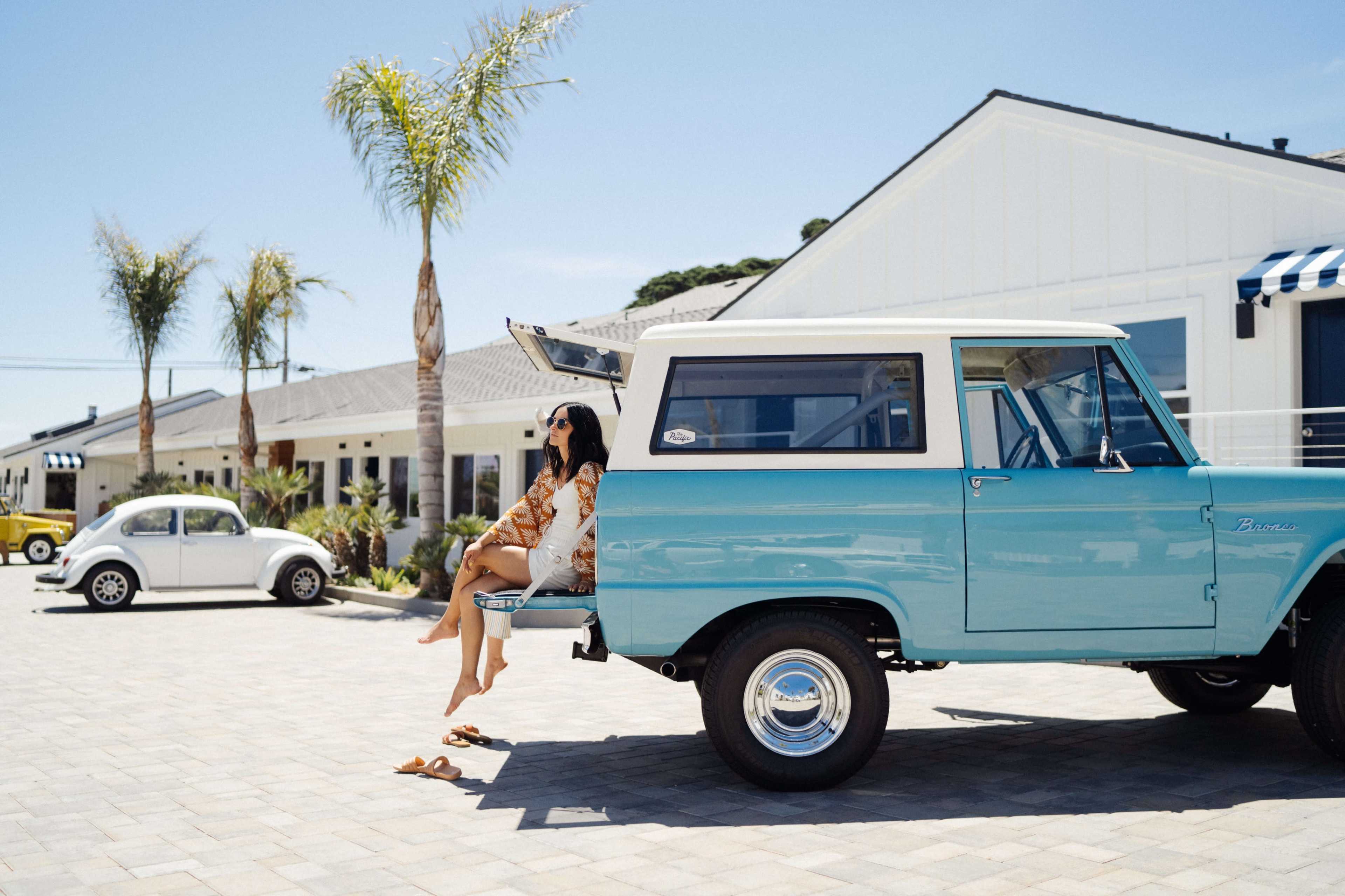 A woman sits on the open tailgate of a blue vintage Bronco parked in a sunny outdoor space with palm trees and nearby cars.