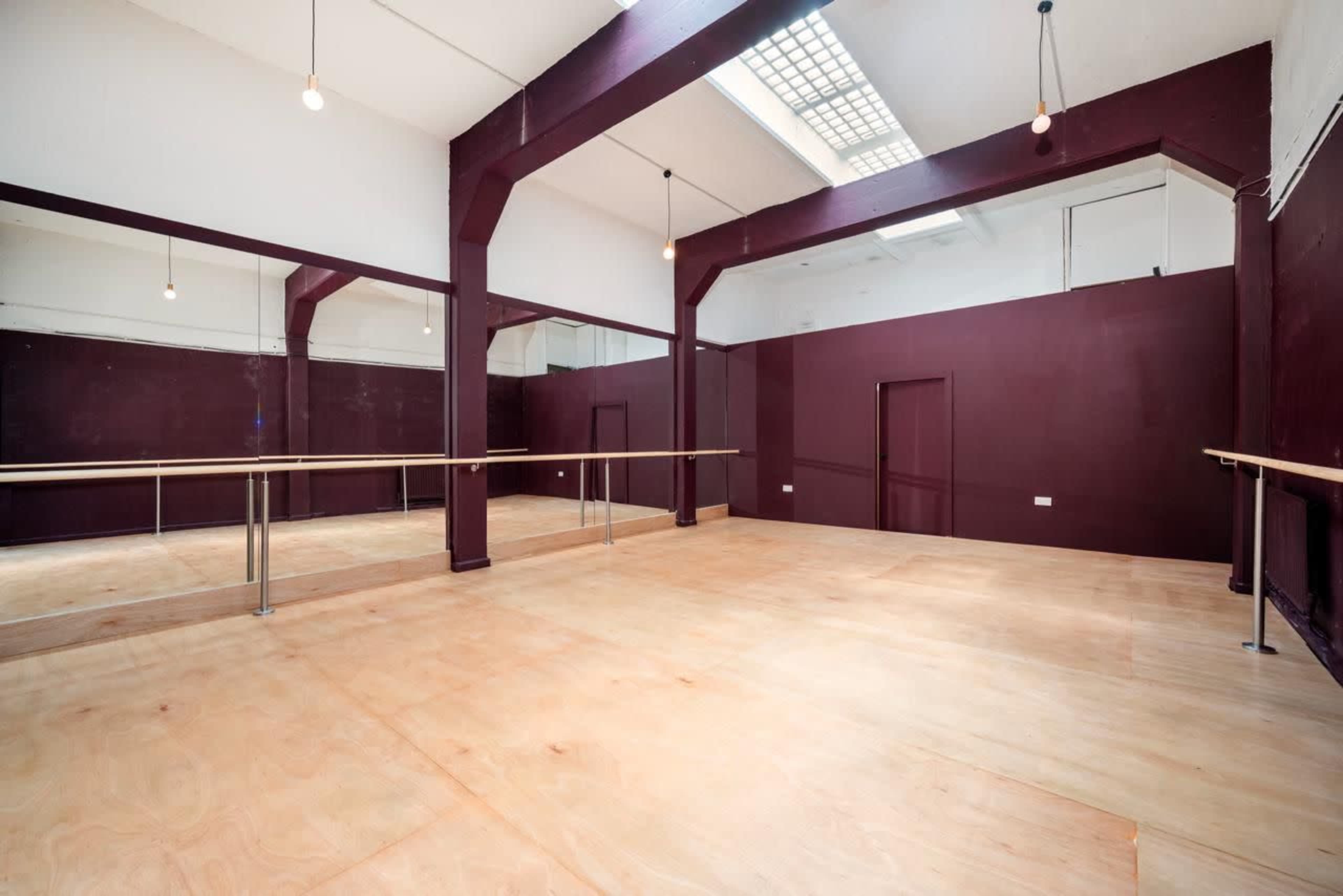 The image shows a spacious dance studio with wooden floors, mirrored walls, and exposed light bulbs hanging from the ceiling.