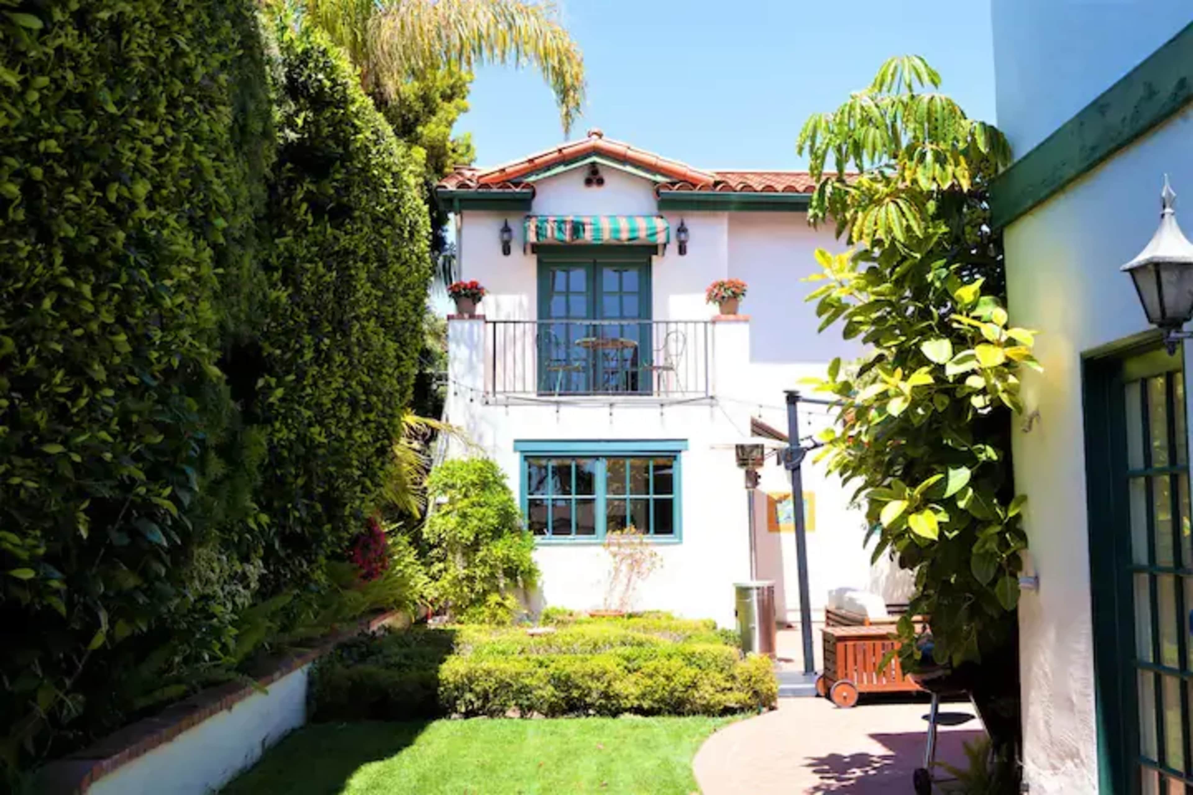 A narrow pathway leads to a two-story house surrounded by green hedges and a well-maintained garden with a patio.