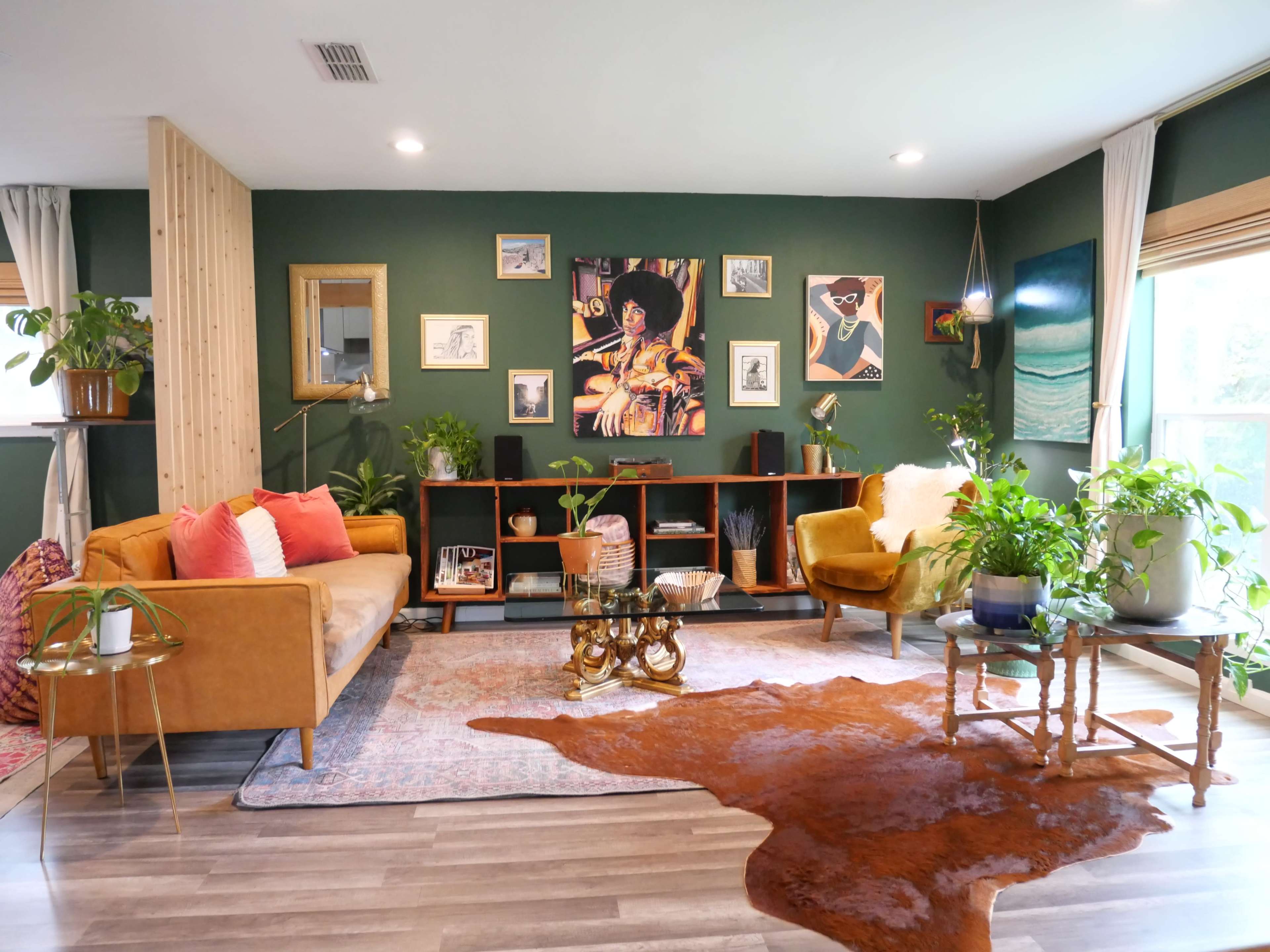 A stylish living room with a green accent wall, a brown sofa, a yellow armchair, a wooden shelf with art and plants, and a rug on the floor.
