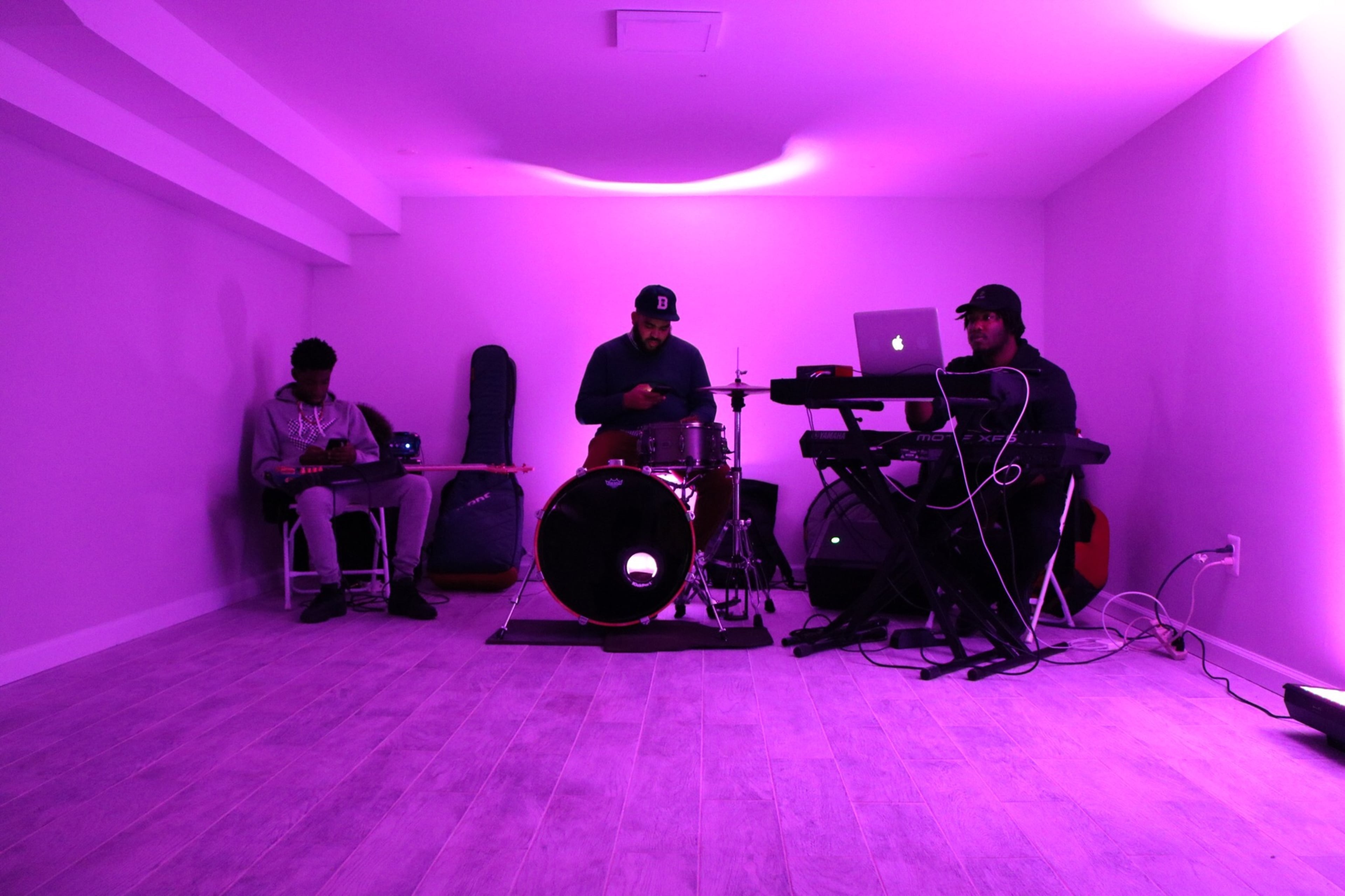 Three musicians are performing in a dimly lit room with purple lighting, featuring a guitarist, a drummer, and a keyboardist.