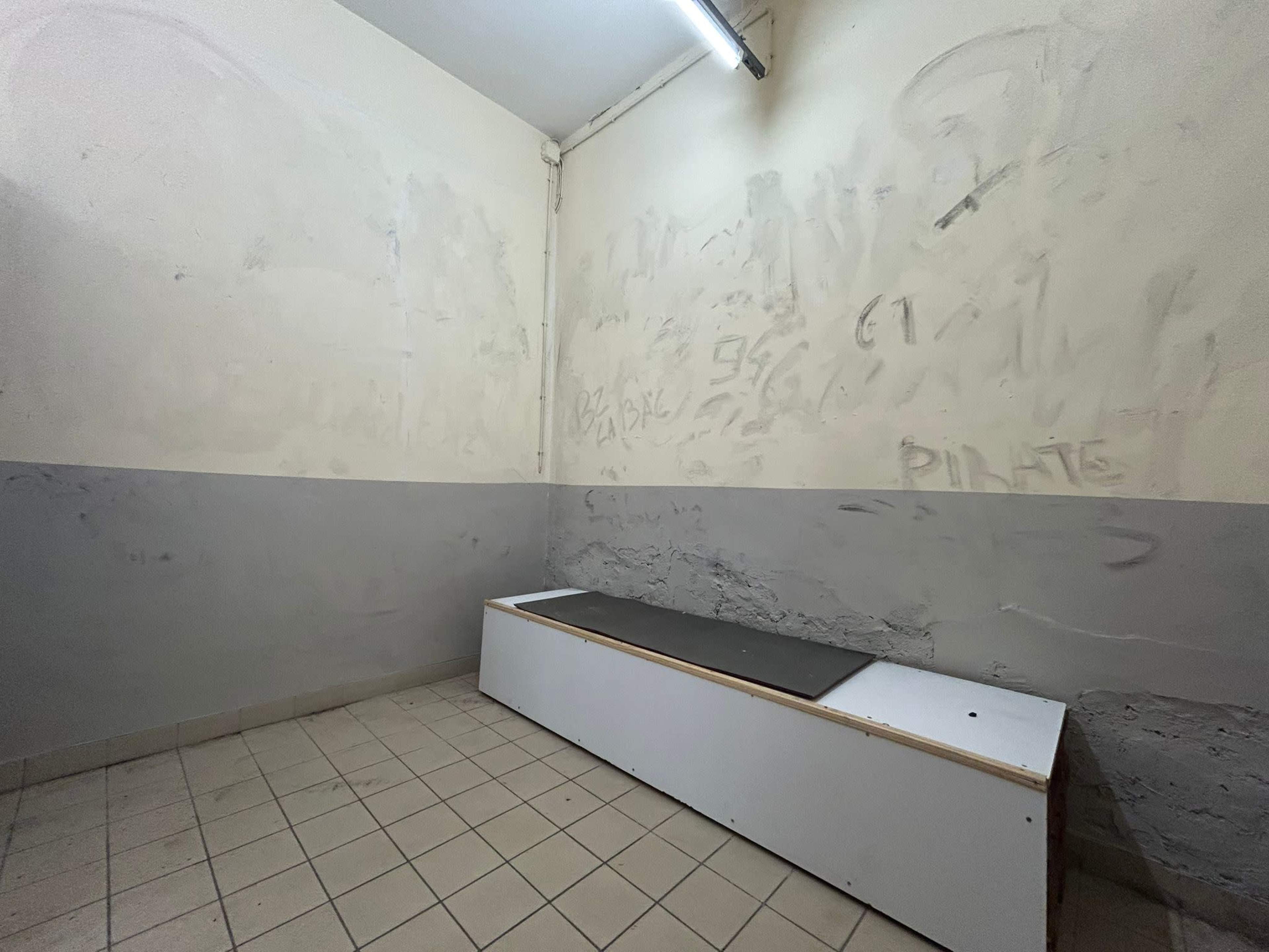 A small, stark room with a plain horizontal platform against one wall and bare, unpainted walls.