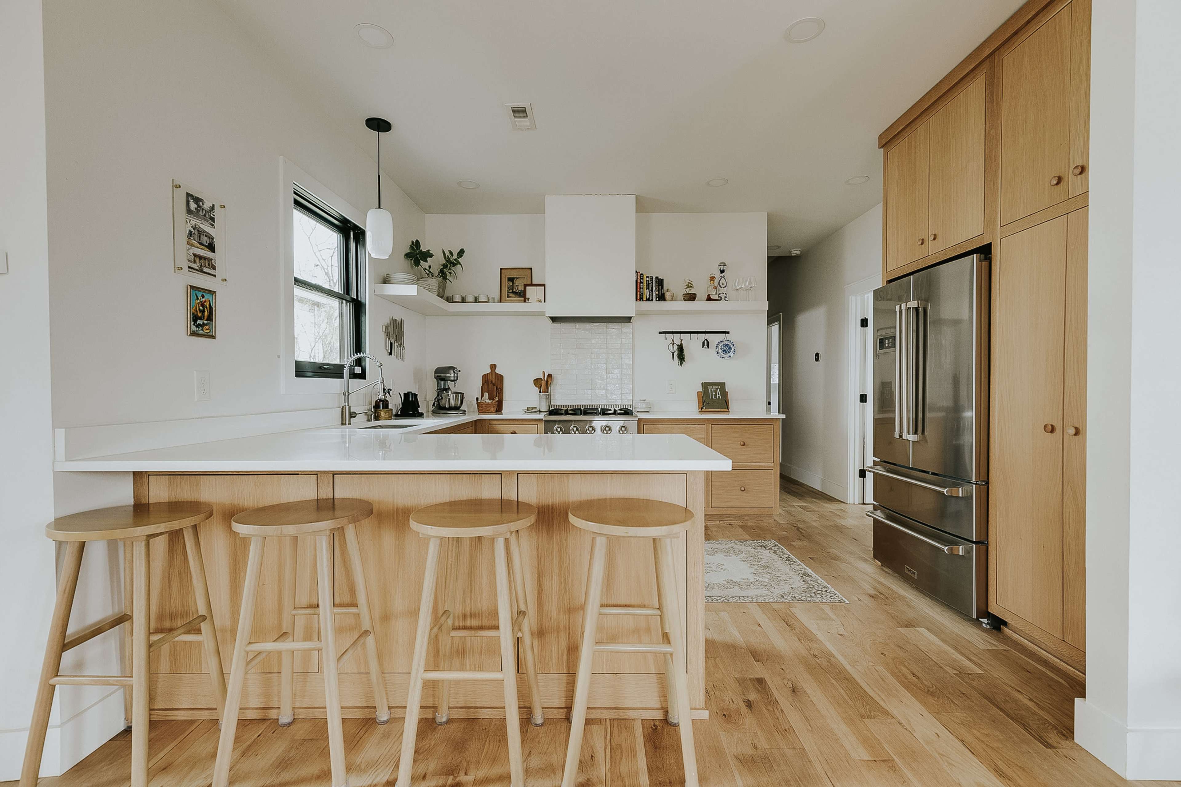 A modern kitchen features a large island with four wooden stools, light wood cabinetry, and stainless steel appliances.