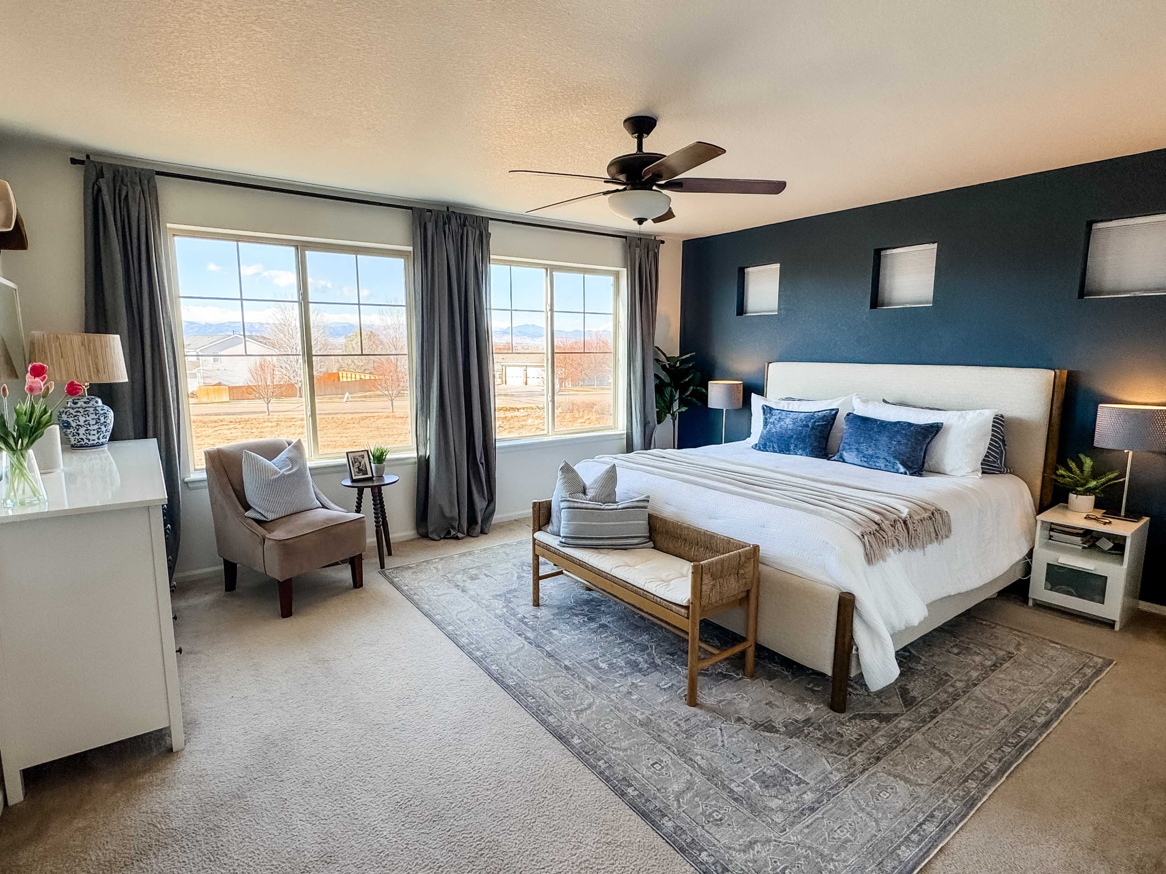 The image shows a neatly arranged bedroom featuring a bed with decorative pillows, a small seating area, and large windows that let in natural light.