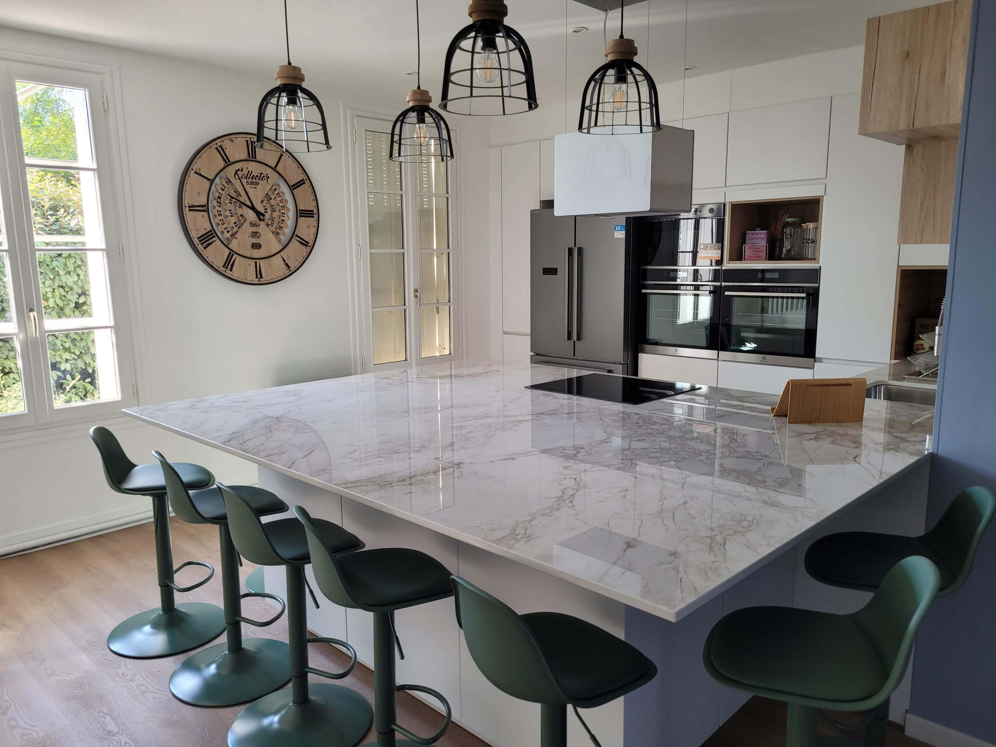 A modern kitchen with a marble island, green bar stools, and pendant lights, featuring a wall clock and built-in appliances.