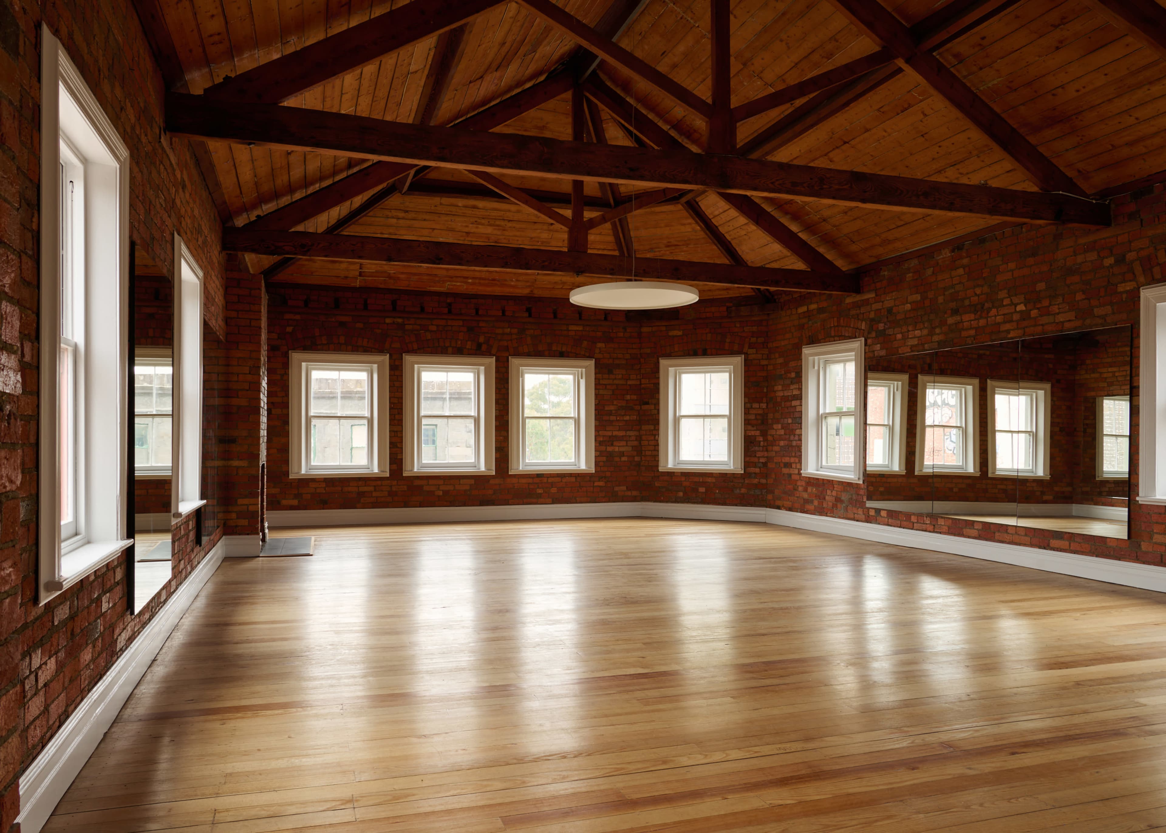 The image shows a spacious room with wooden flooring and exposed brick walls, featuring several windows and a high, beamed ceiling.