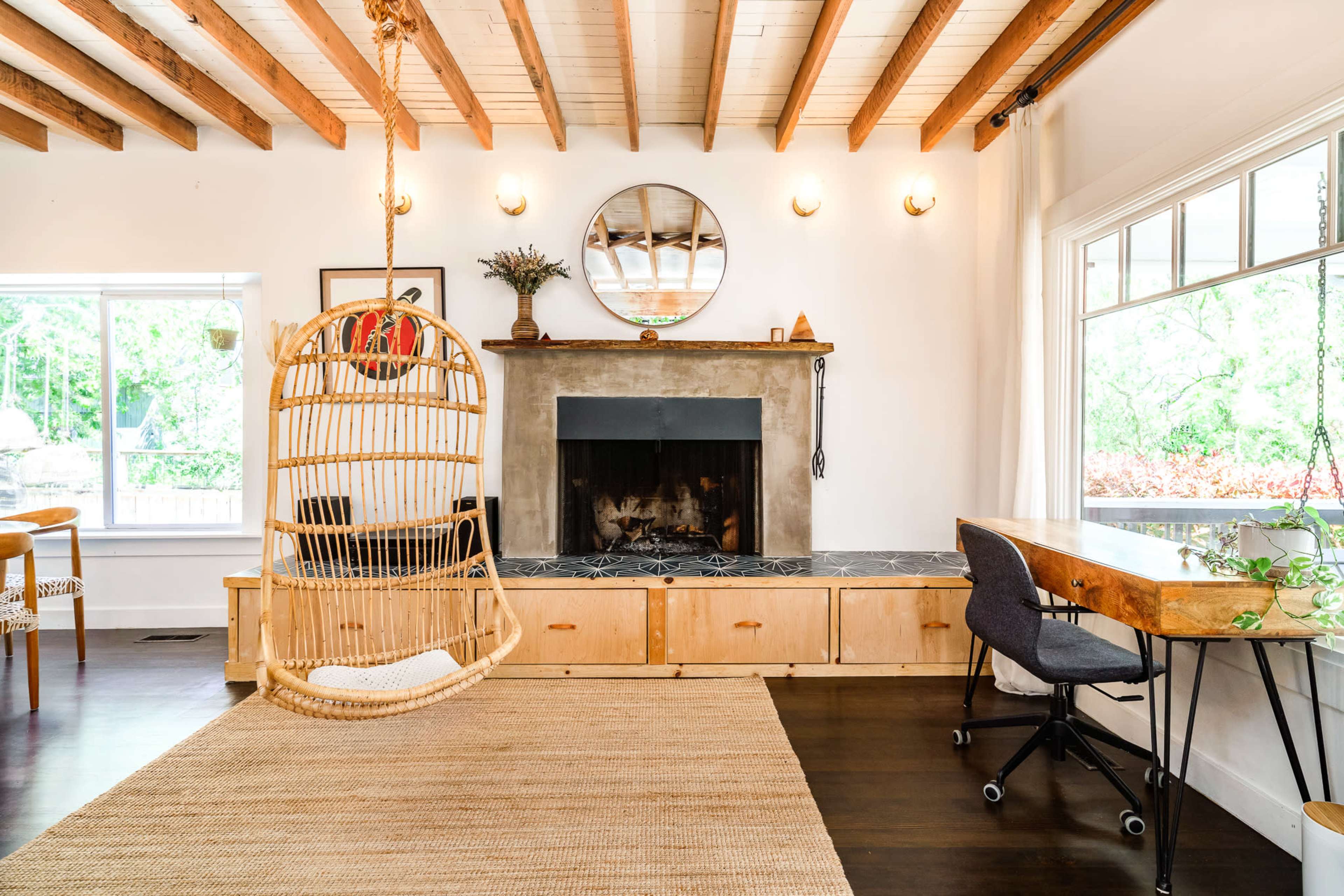 The image depicts a modern living room featuring a hanging rattan chair, a stone fireplace, and a wooden table beside a window.