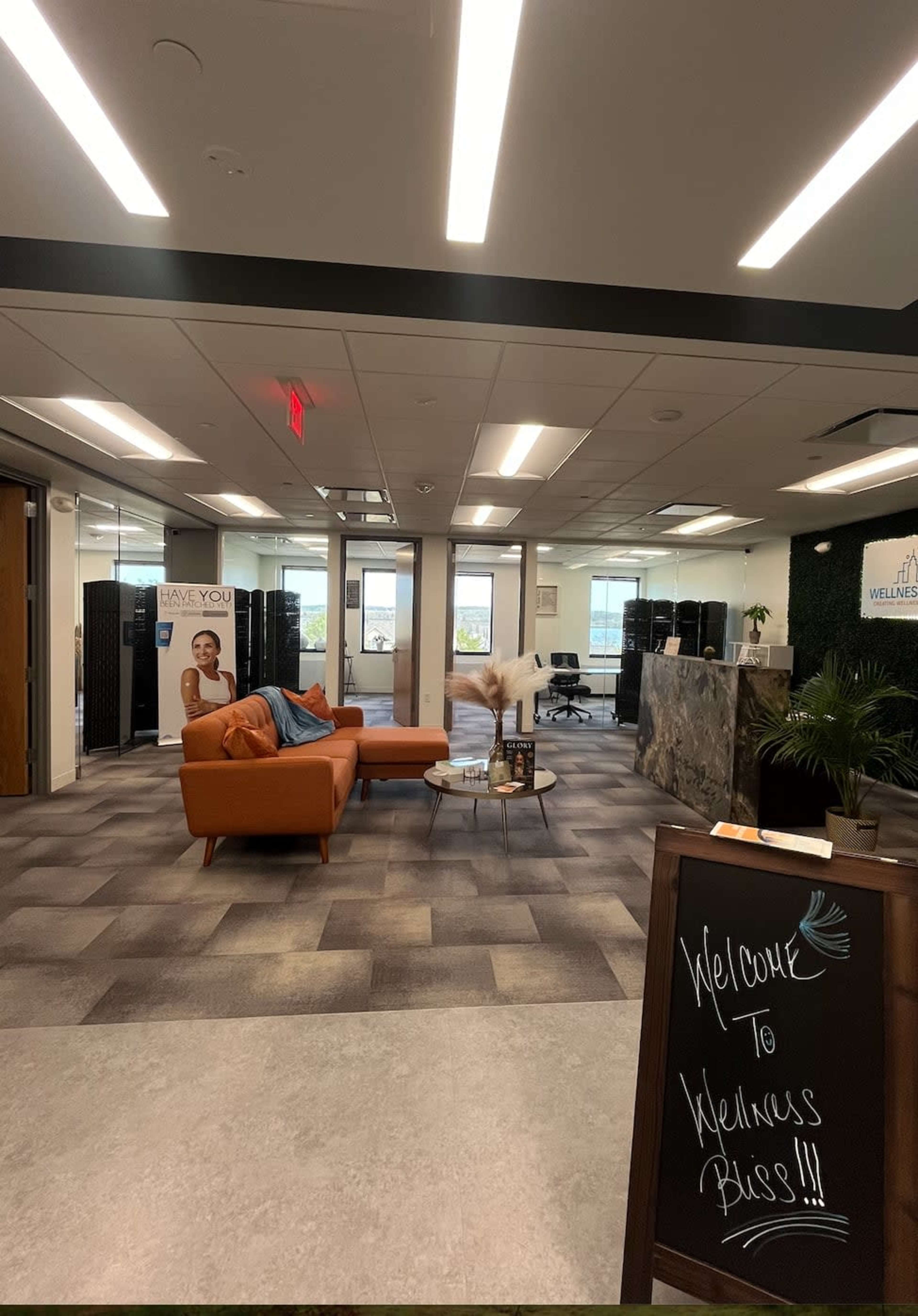 A modern office reception area with a seating arrangement featuring an orange couch, a welcome sign, and large windows offering a view outside.