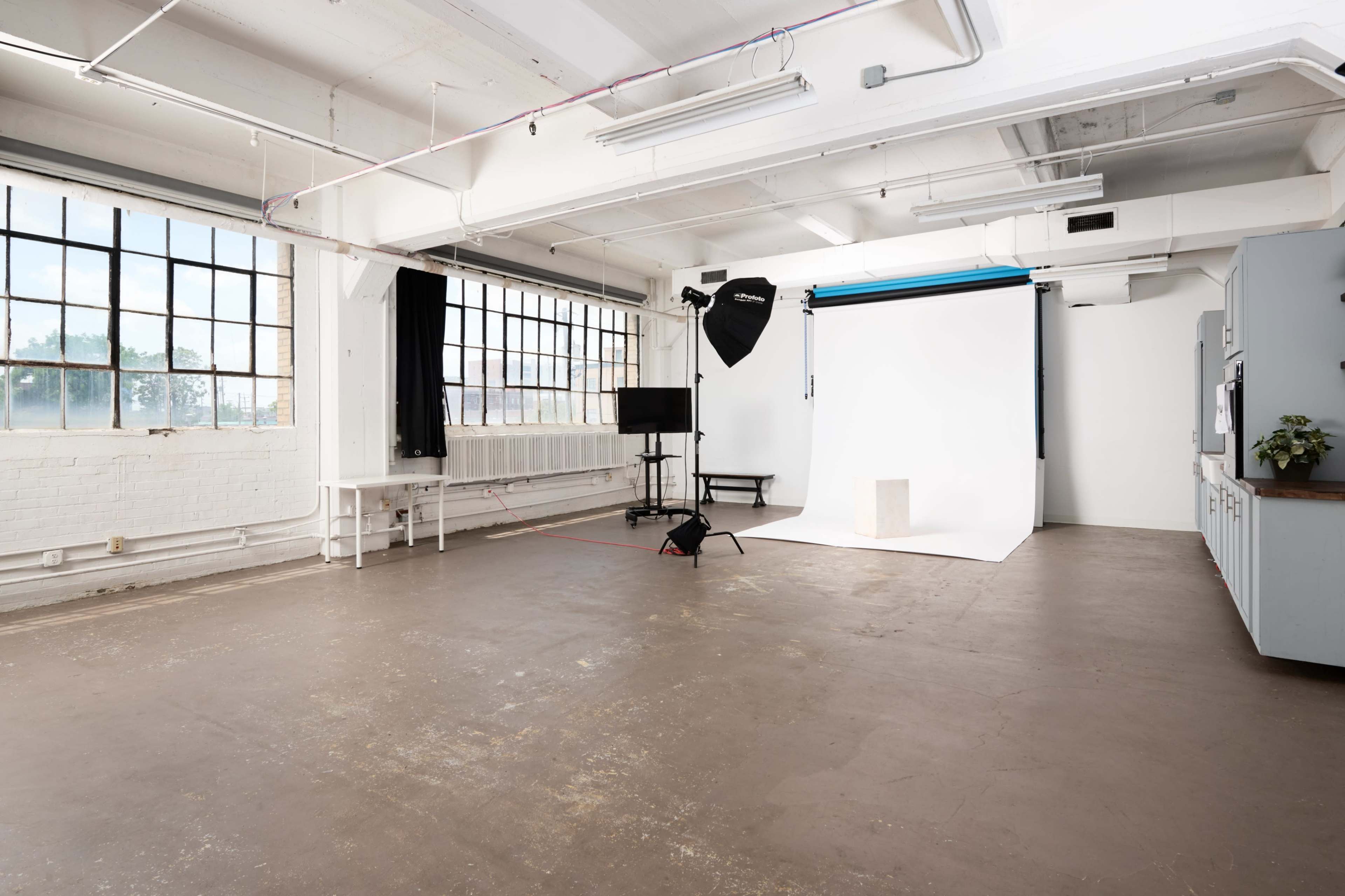 The image shows a spacious, well-lit photography studio with large windows, a white backdrop, and equipment set up on the floor.