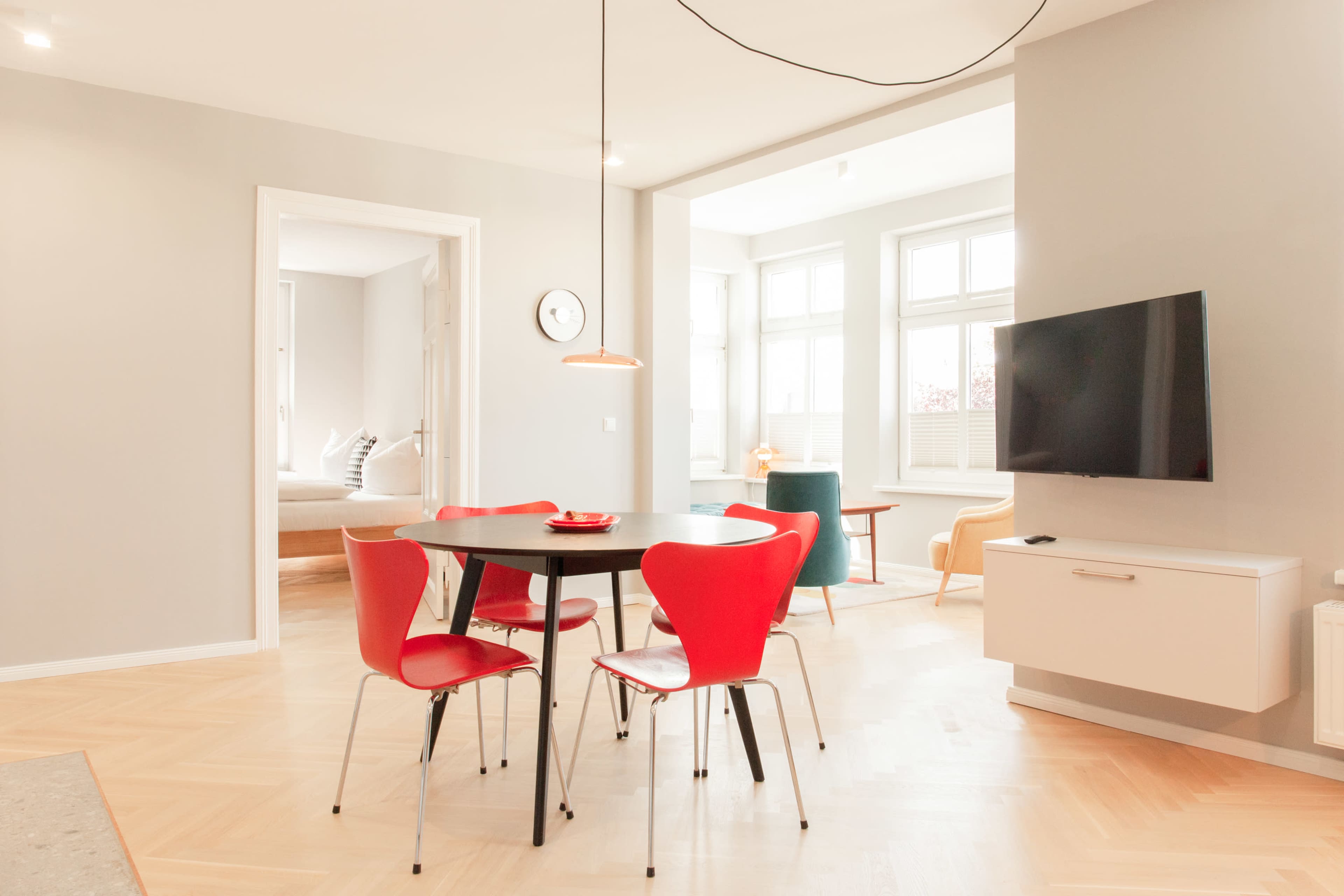 A dining area features a black circular table surrounded by four red chairs, with a modern light fixture overhead and a television mounted on the wall.