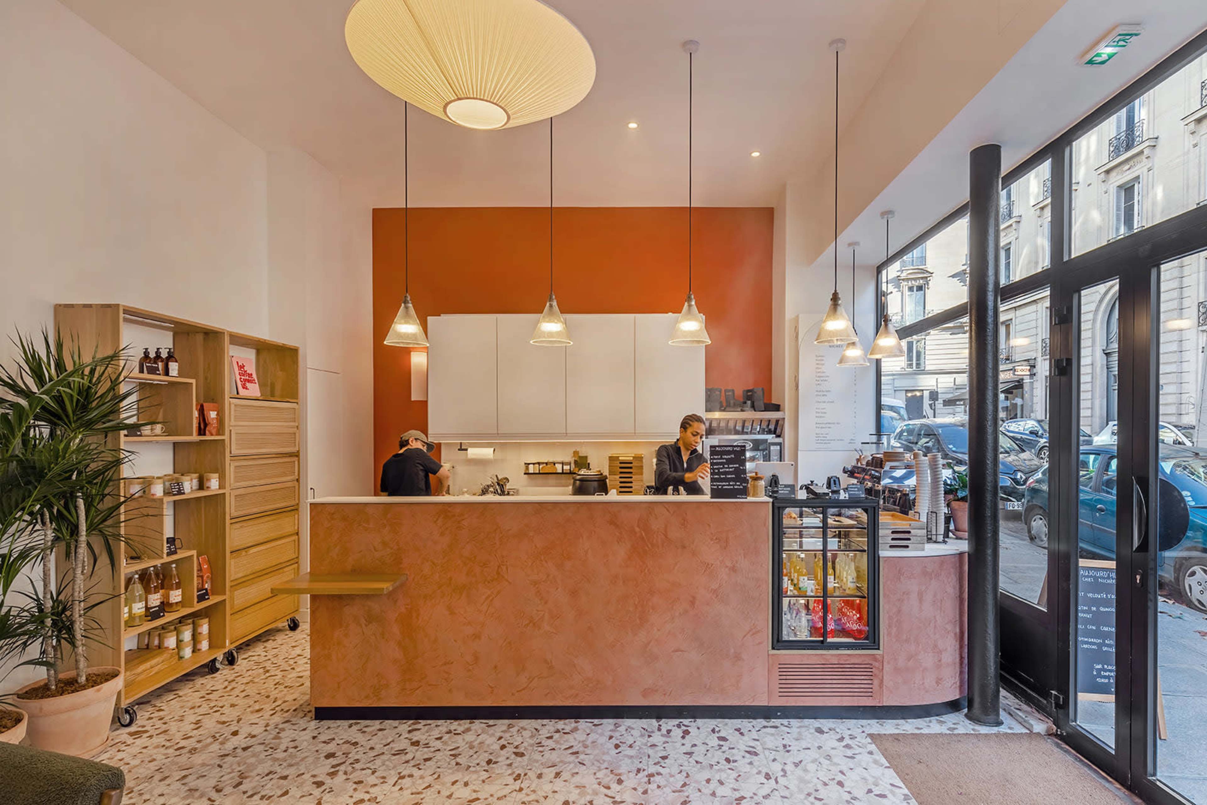 The image shows a modern coffee shop interior with a light-colored counter, pendant lighting, and a display refrigerator, featuring two staff members working behind the counter.
