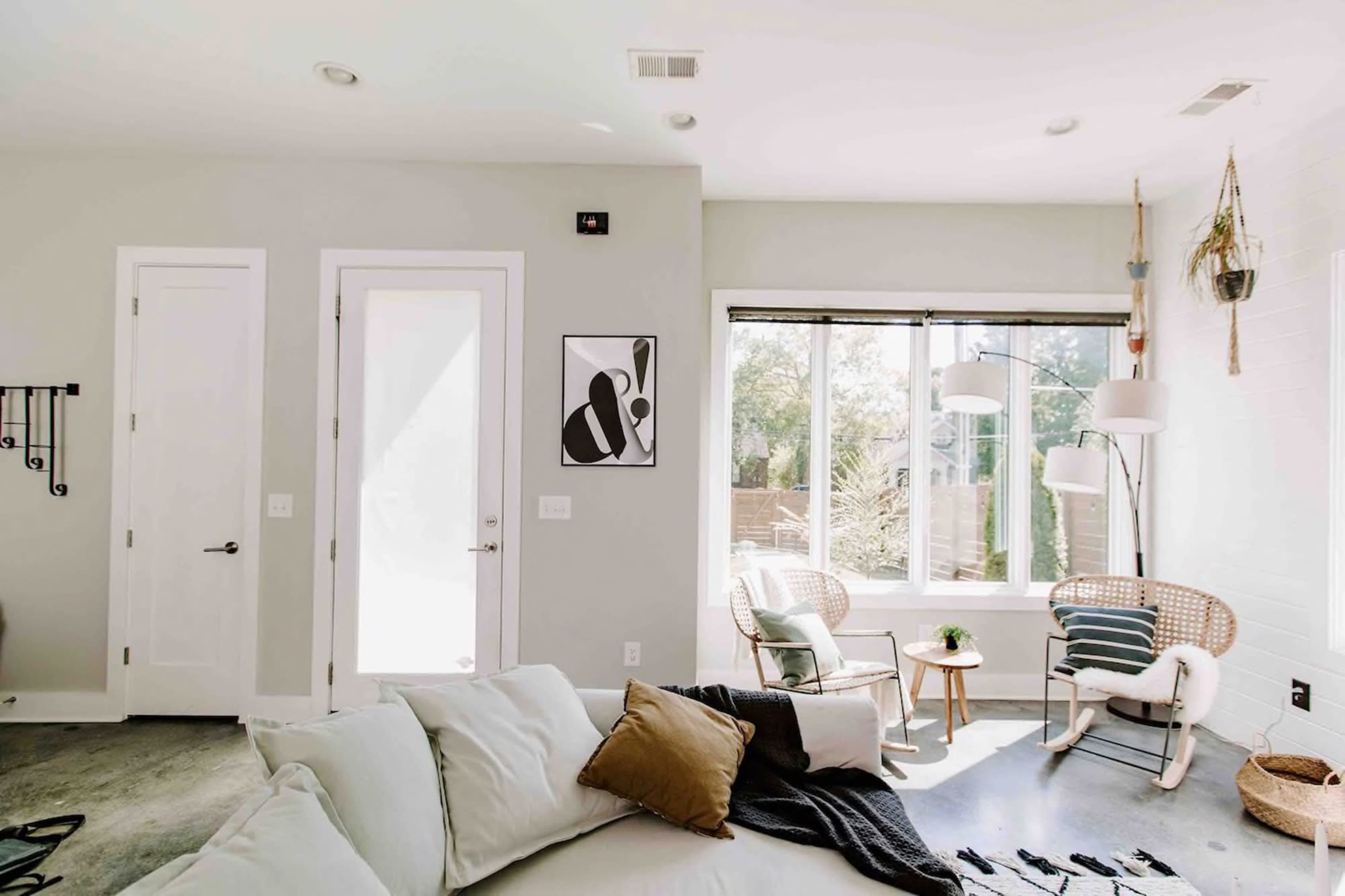 A modern living room with a large window, a light-colored sofa, a door, and two chairs near the window.