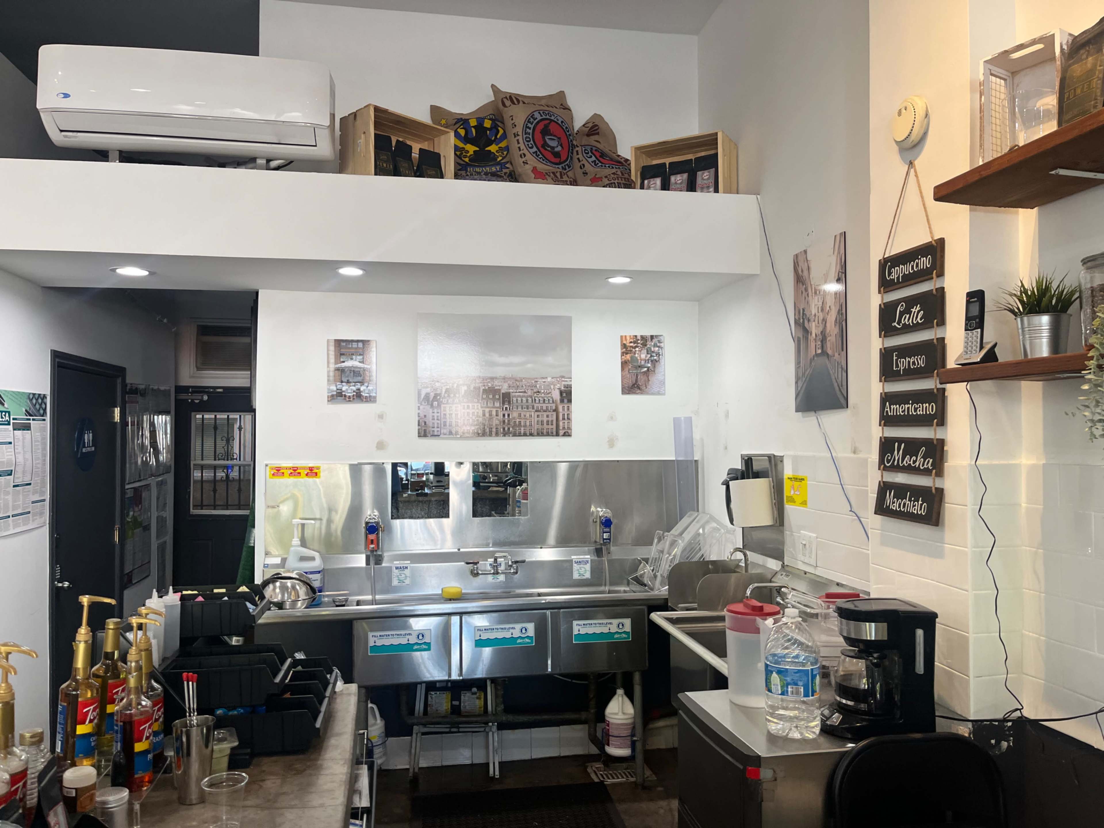 The image shows a small coffee shop kitchen with equipment, a sink, and various supplies organized on shelves.