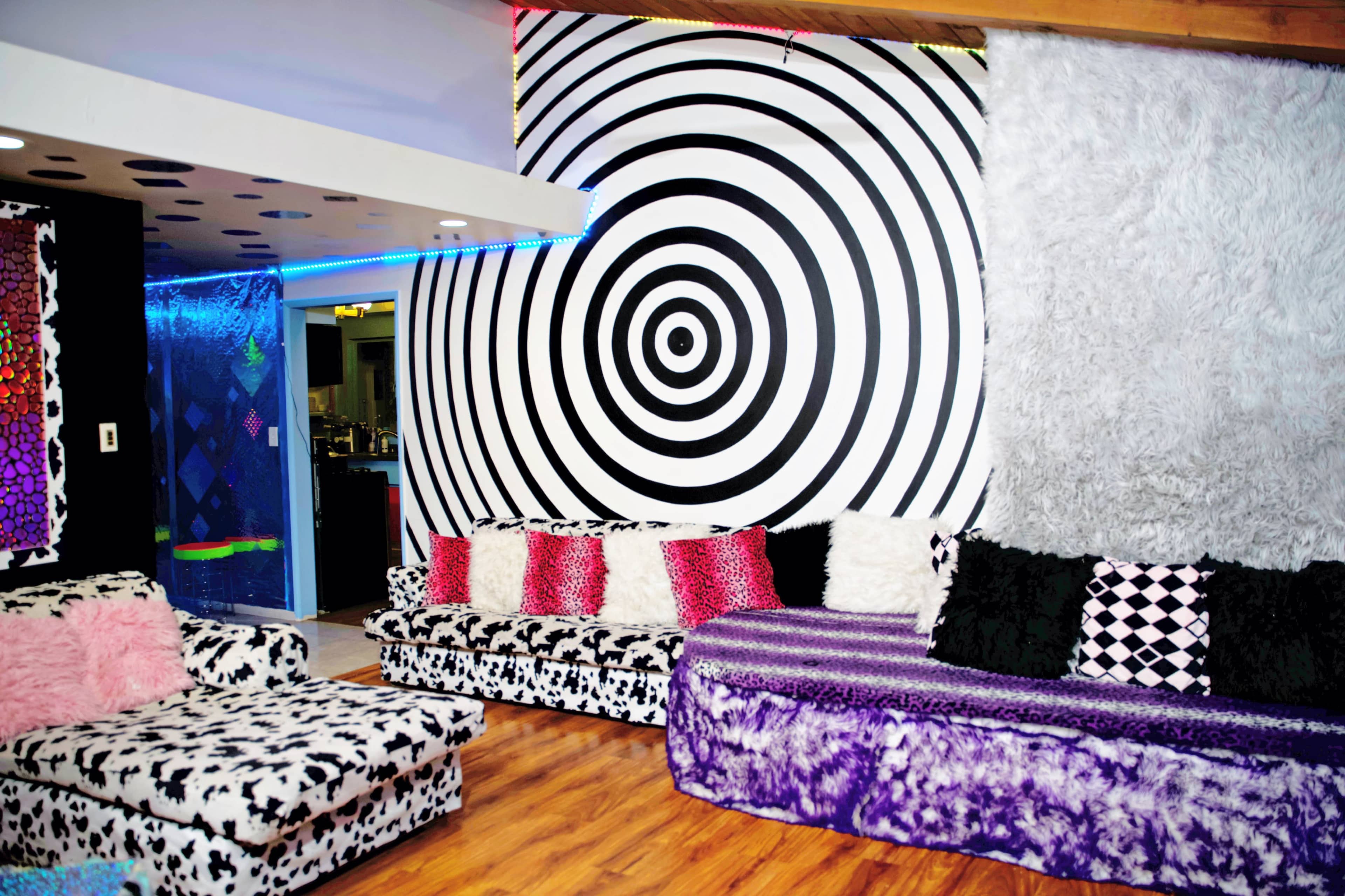 The room features black and white spiral patterns on the wall, colorful decorative couches, and a wooden floor.