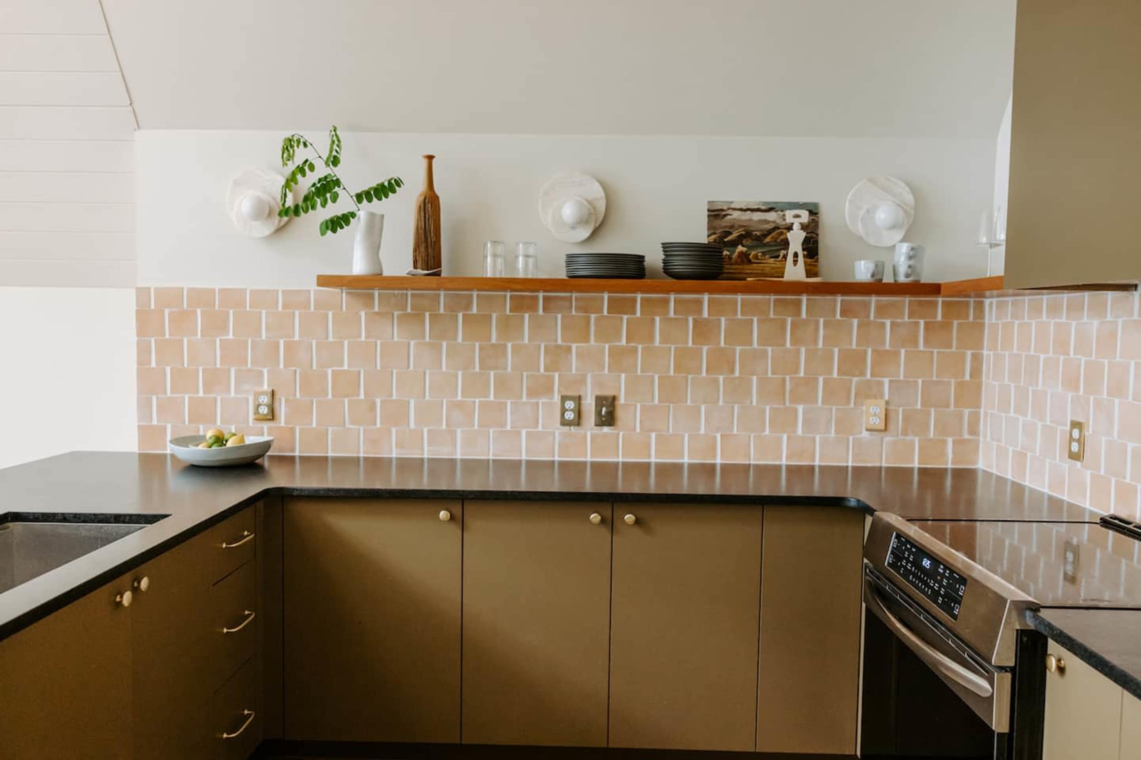 A modern kitchen features a beige tiled backsplash, dark countertops, and open shelving displaying ceramics and glassware.