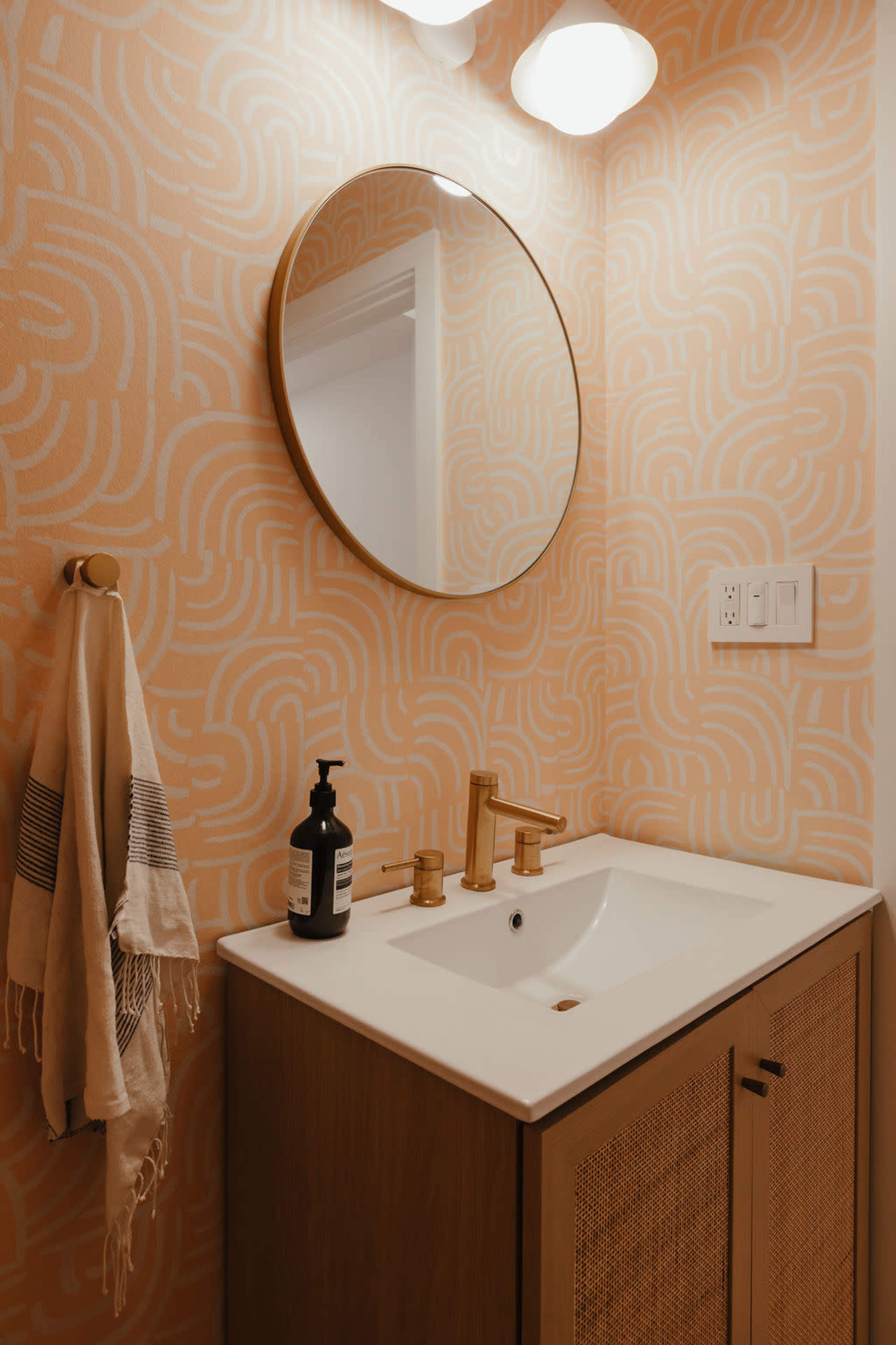 The image shows a modern bathroom vanity with a round mirror, a white sink, gold faucet fixtures, and patterned peach wallpaper.