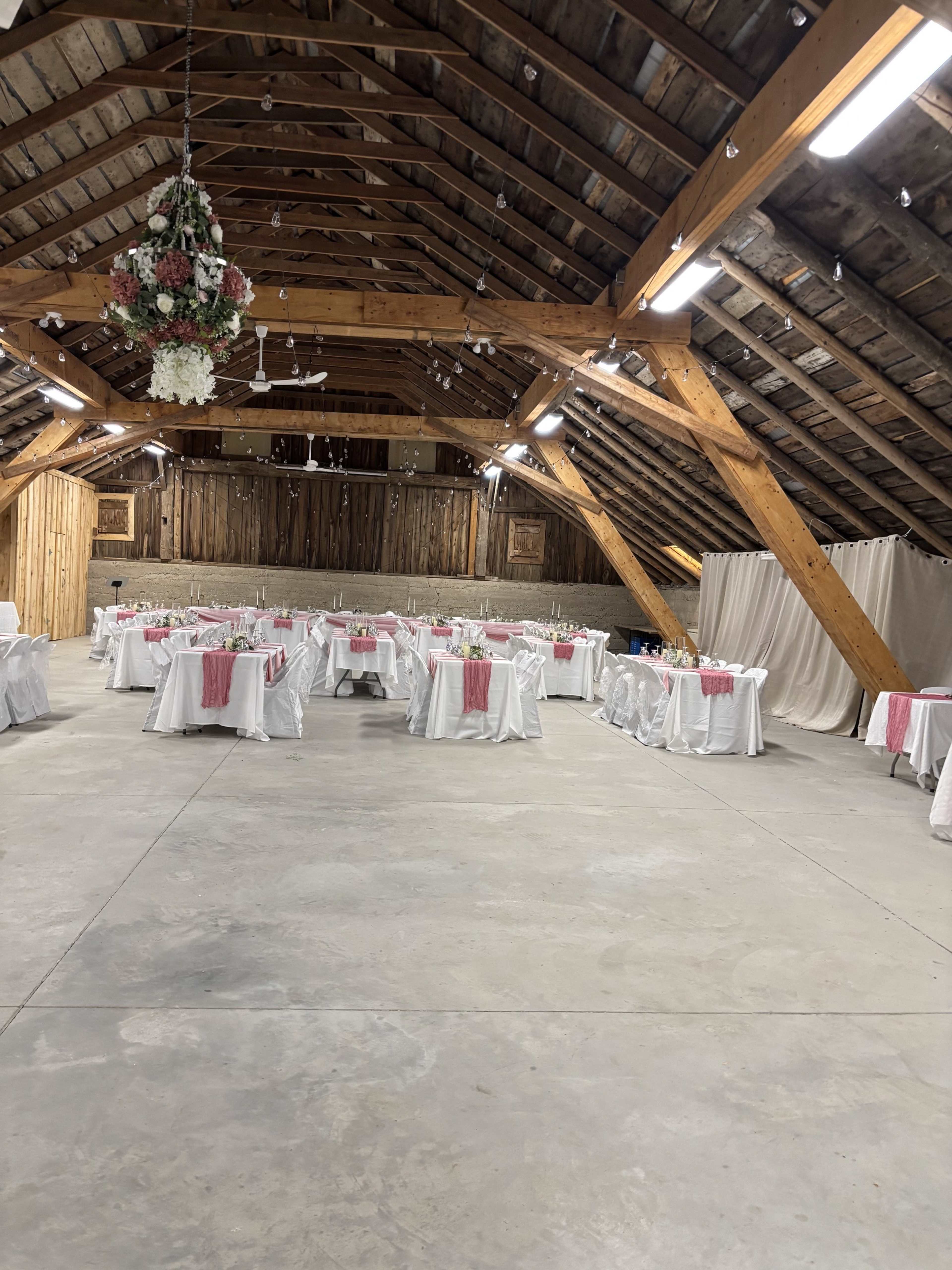 The image shows a spacious barn with wooden beams, featuring several tables covered in white tablecloths with pink accents arranged for an event.