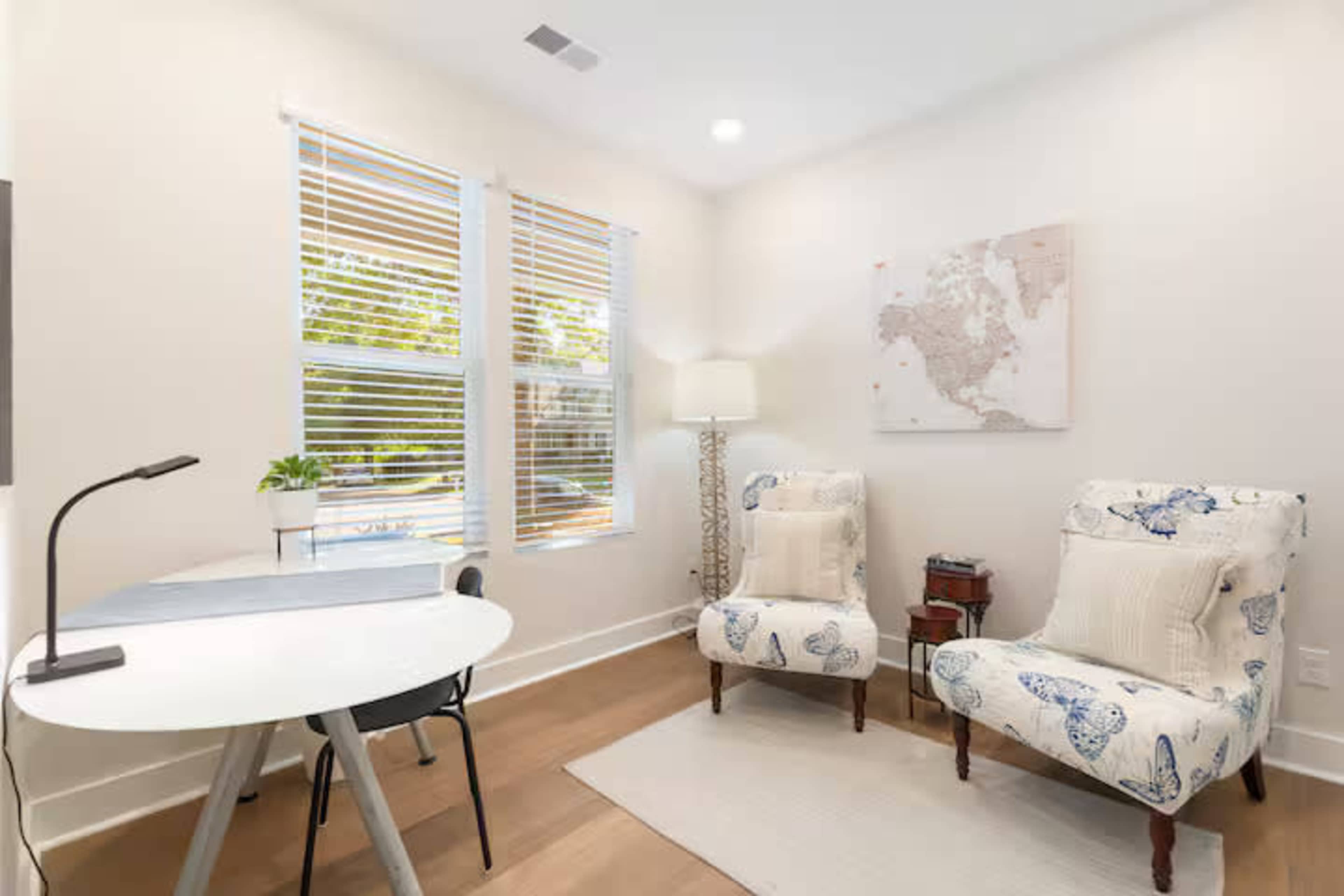 A bright room features two patterned armchairs, a small round table, and a lamp by a window with blinds, alongside a world map on the wall.