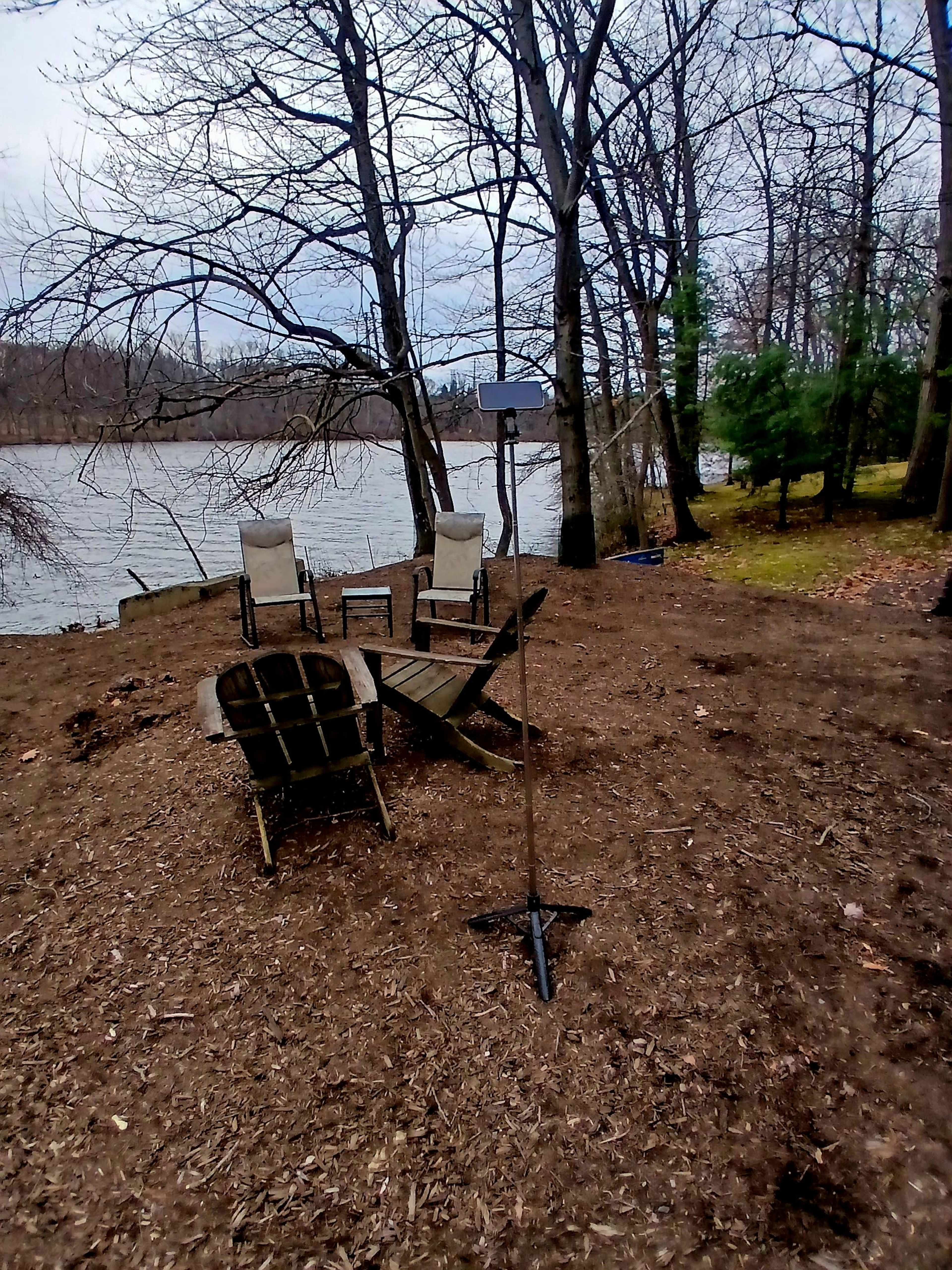 A clearing near the water features three chairs arranged in a circle, with a small table and a pole in the center, surrounded by trees.