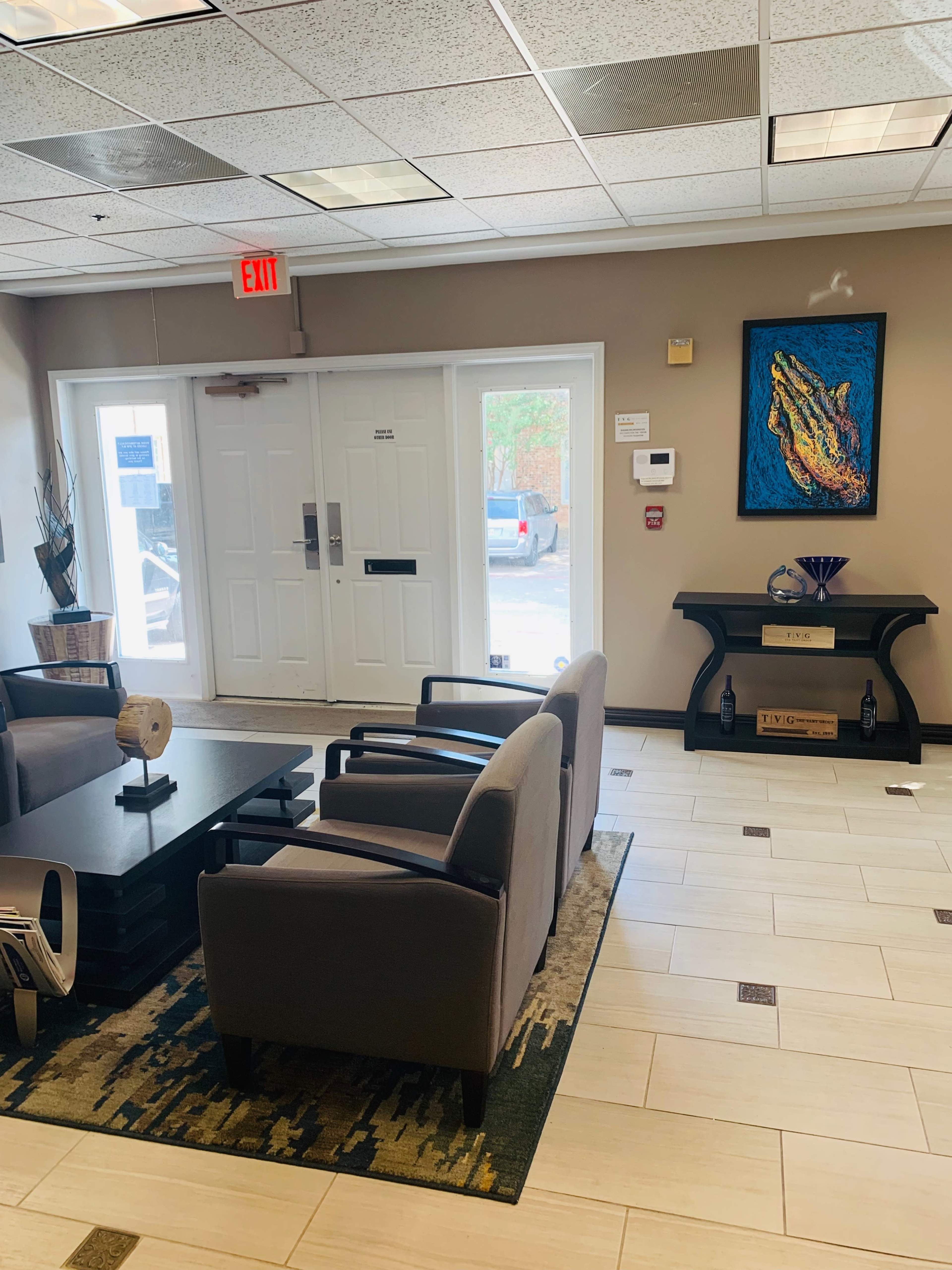 The image shows a waiting area featuring four chairs around a coffee table, with an art piece of an alligator on the wall, and a door leading outside.