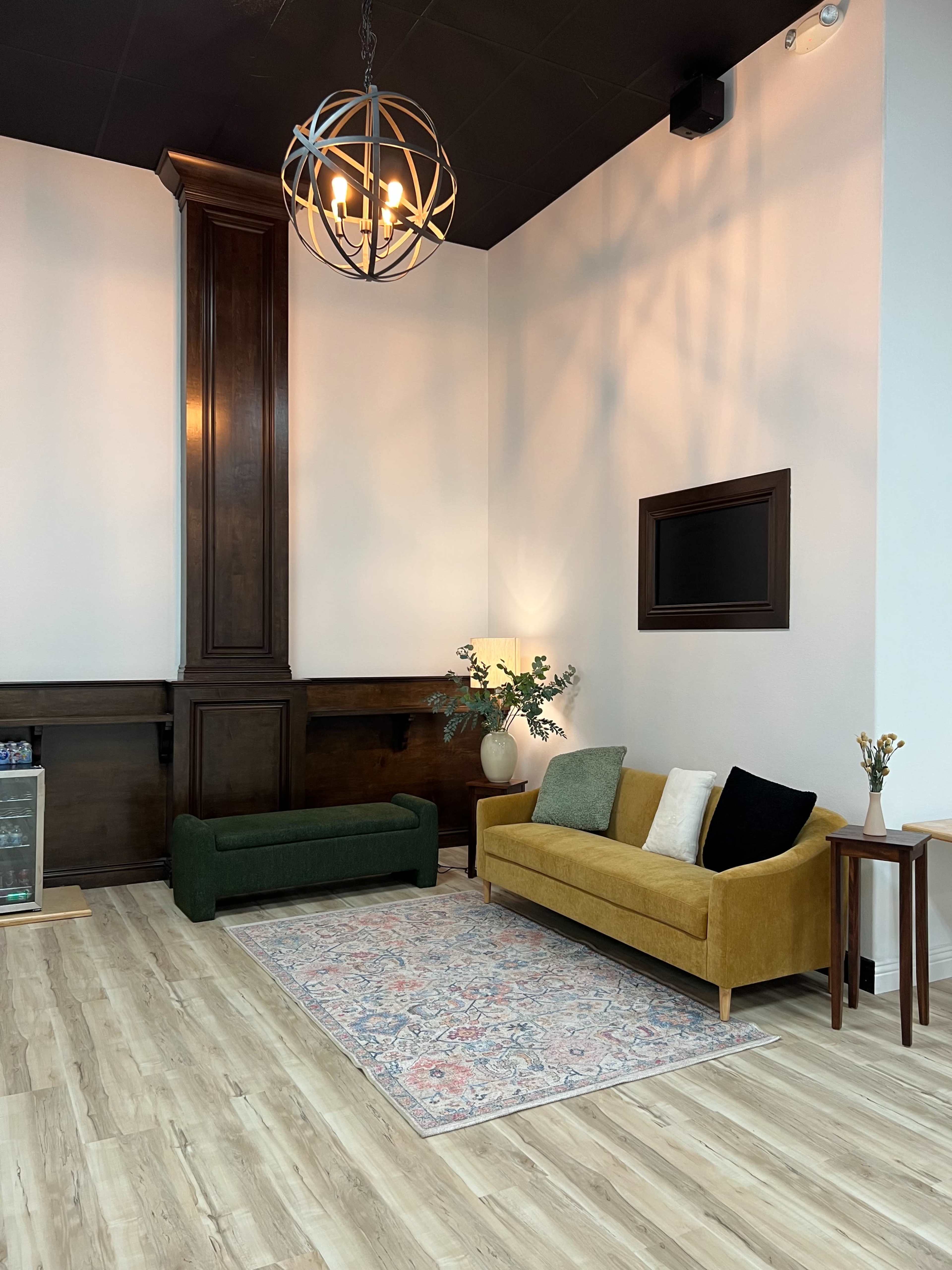 The image shows a modern interior space featuring a yellow sofa, a green bench, a decorative rug, and a plant beside a lamp against a light-colored wall.