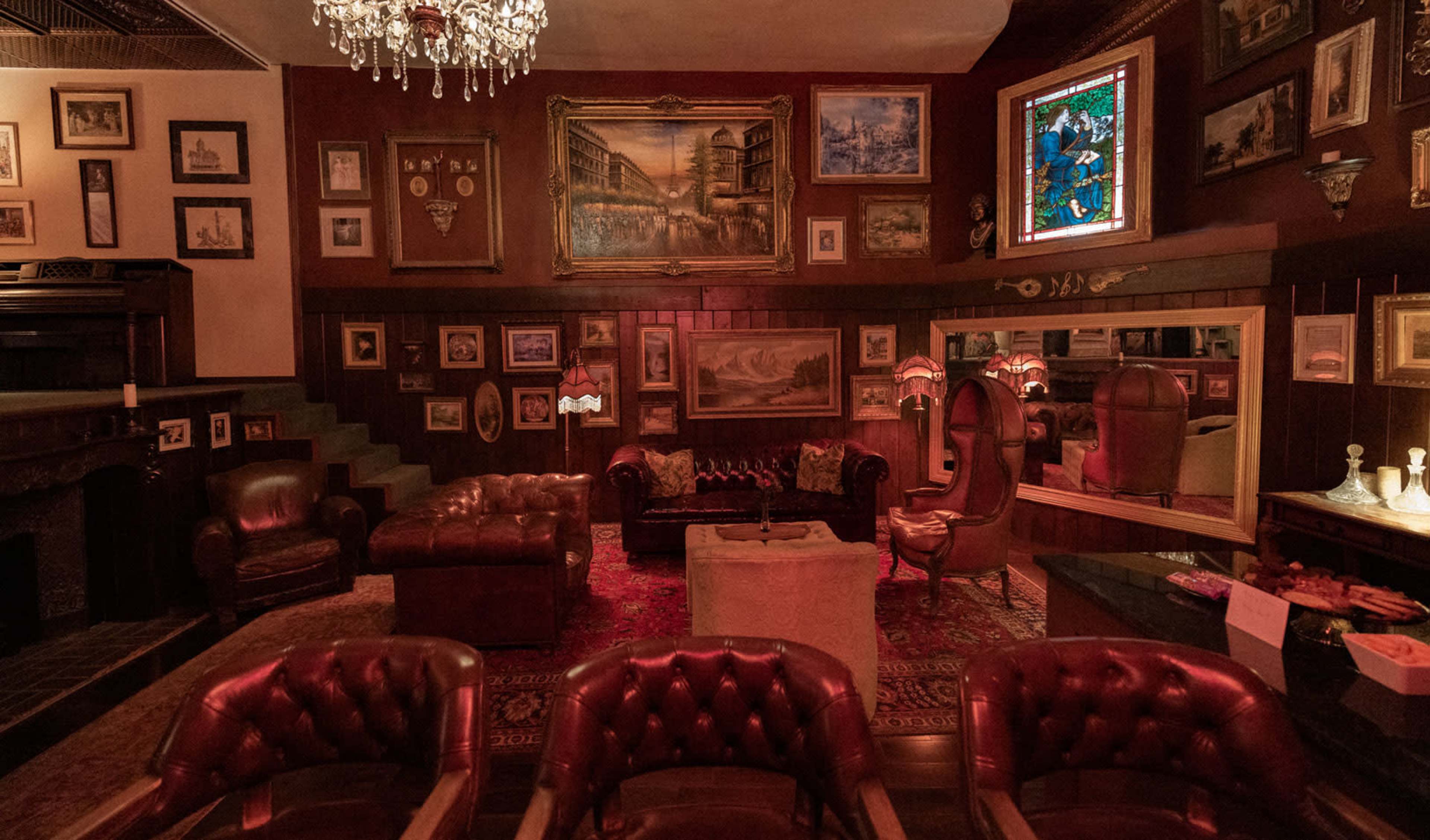 The image shows a dimly lit, vintage-style lounge decorated with an array of framed artworks, ornate furniture, and a chandelier.