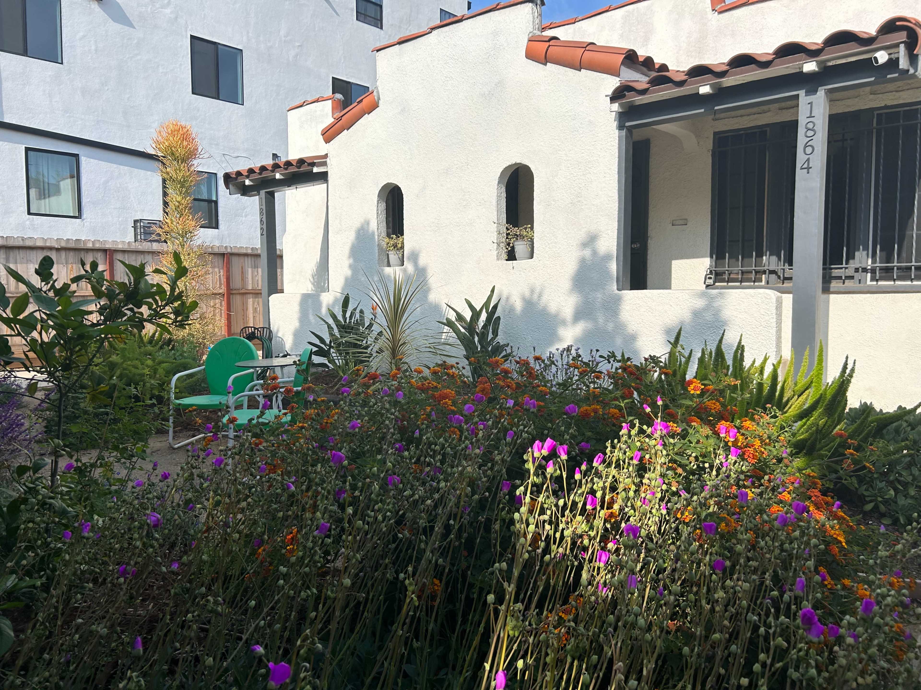 A small garden filled with vibrant flowers and plants surrounds a white stucco building with a tiled roof and visible windows.