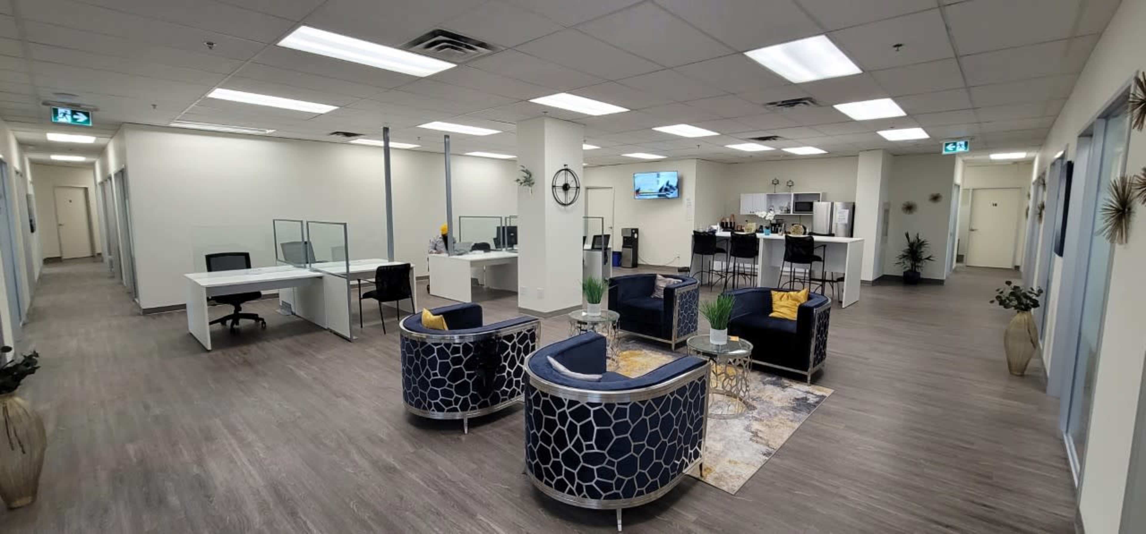 A modern office space with several workstations, a lounge area featuring two chairs, and a kitchenette visible in the background.