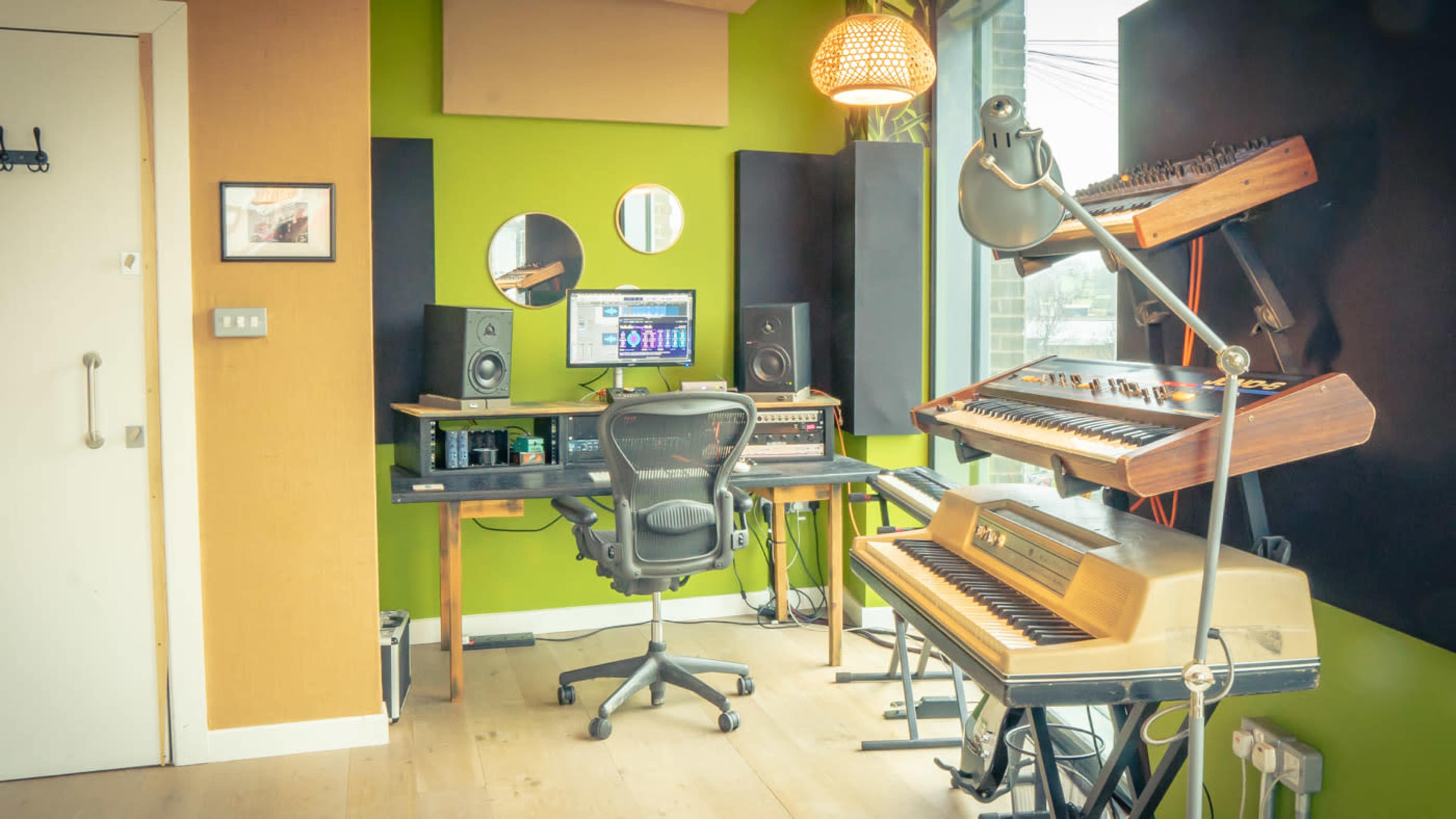 The image shows a music studio with a desk featuring audio equipment, speakers, a computer, and various keyboard instruments against a green and neutral-colored wall.