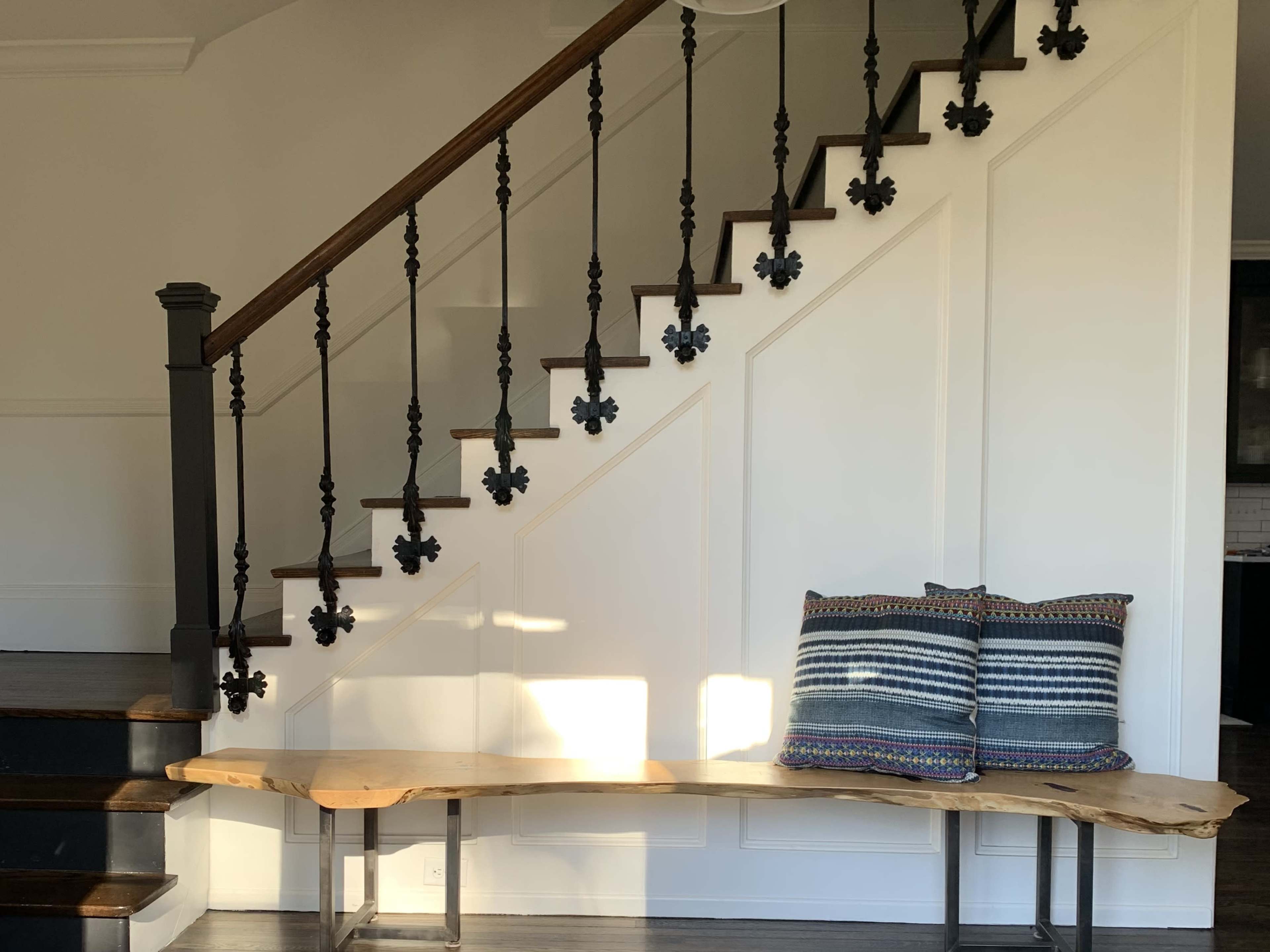 A wooden bench sits against a wall near a staircase with black wrought-iron railings and sunlight streaming across the floor.