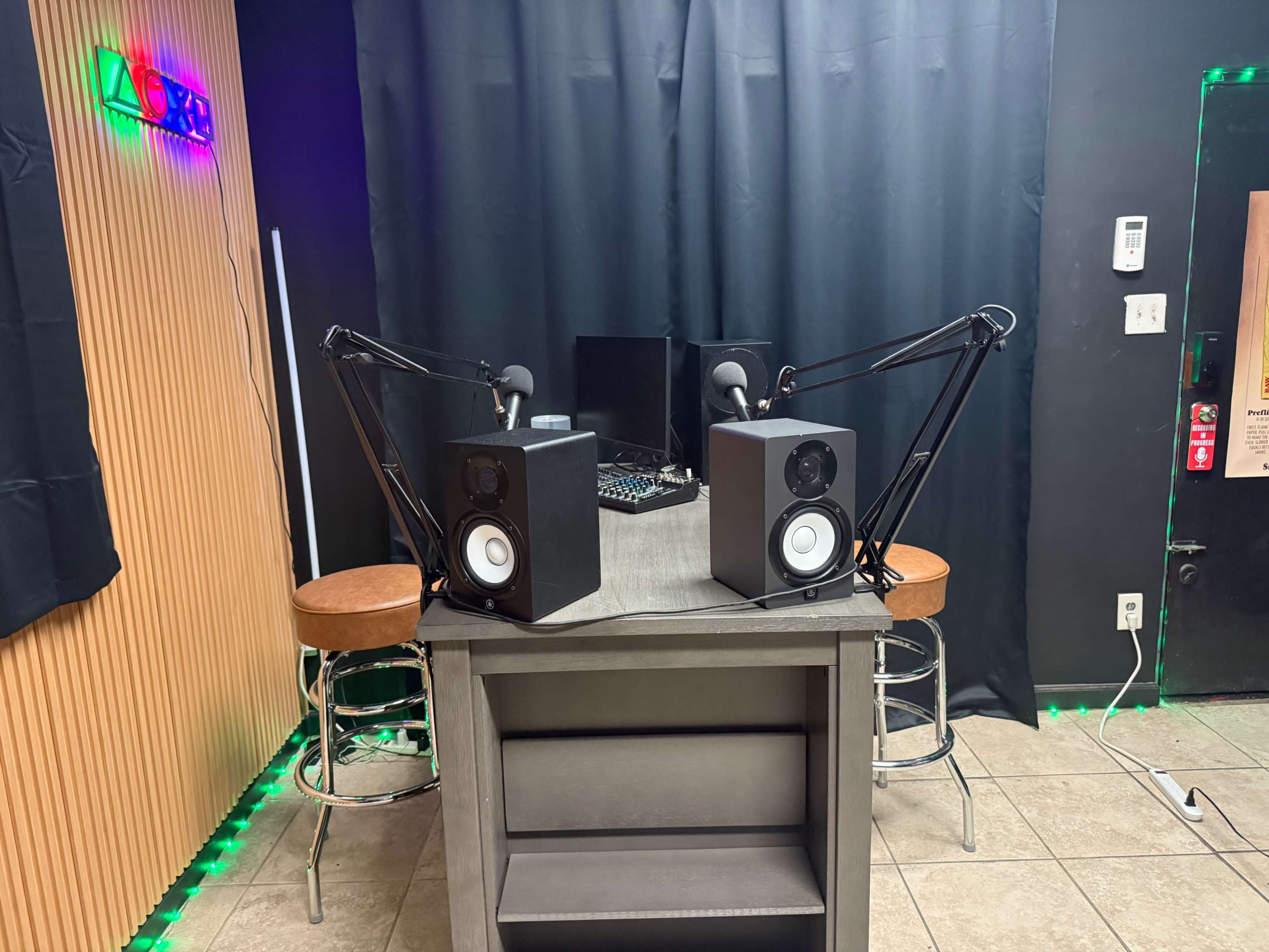 The image shows a recording studio with a table at the center, two microphone stands, and audio speakers, all set against a black backdrop.