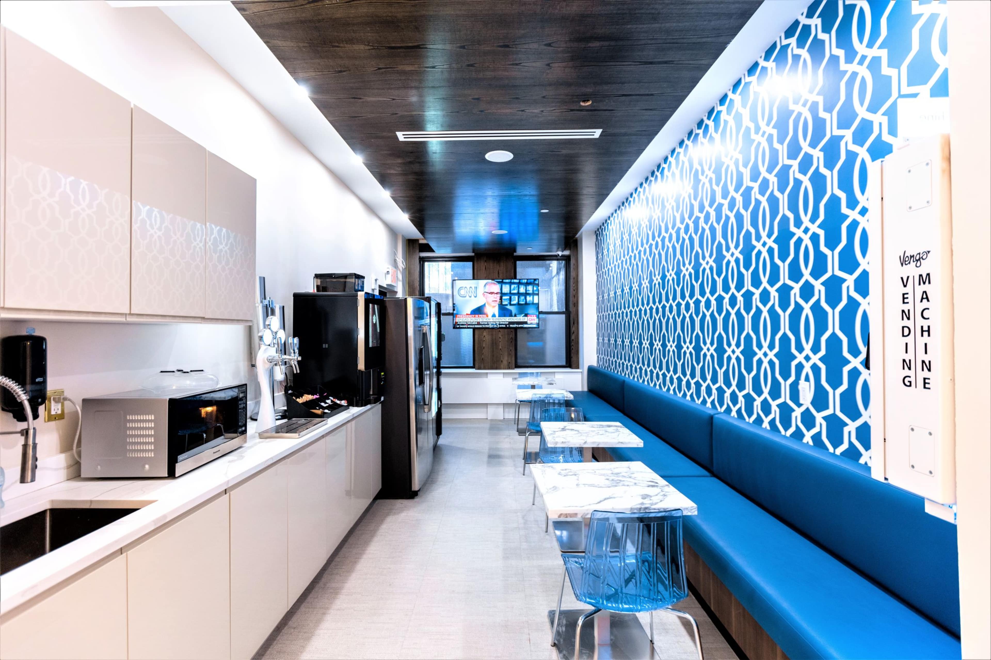 The image shows a modern break room with a kitchen area on one side, featuring a microwave and refrigerator, and a long blue bench along the wall opposite transparent acrylic tables.