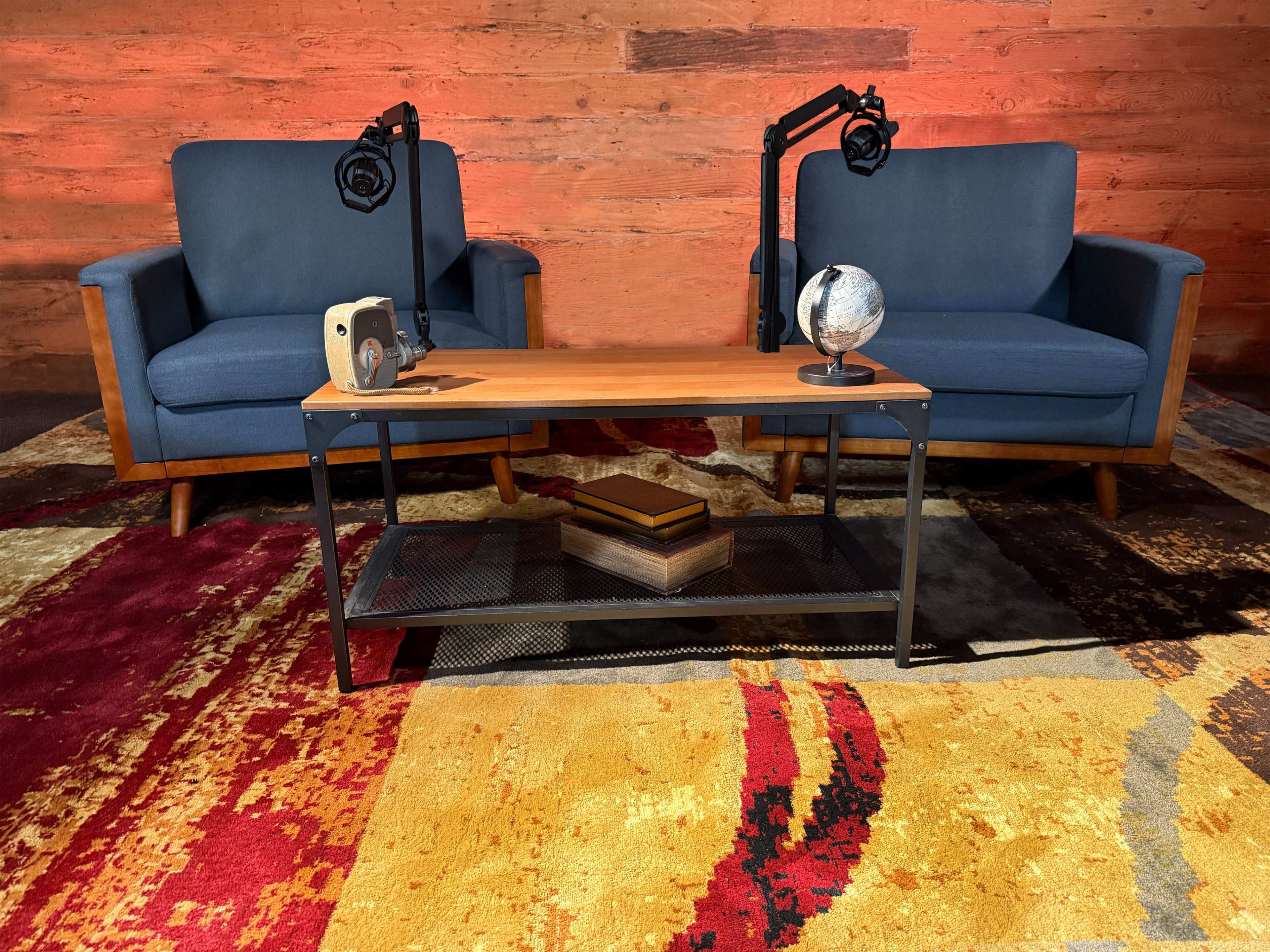 The image depicts a cozy seating arrangement with two blue chairs, a wooden coffee table, and a decorative globe, set against a textured, colorful rug and wooden wall.