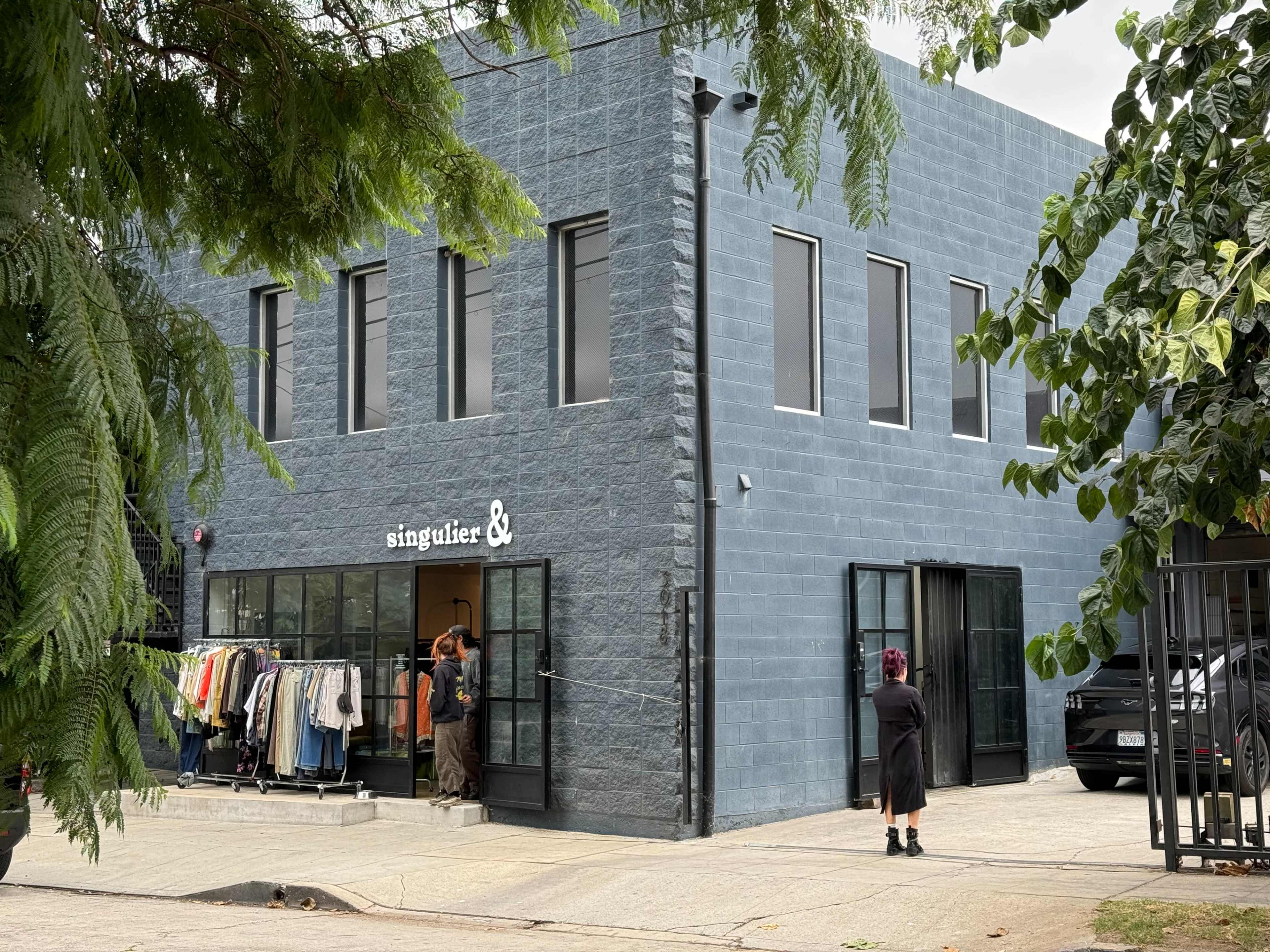 The image shows a gray building with large glass doors, featuring a clothing display outside and two people near the entrance.