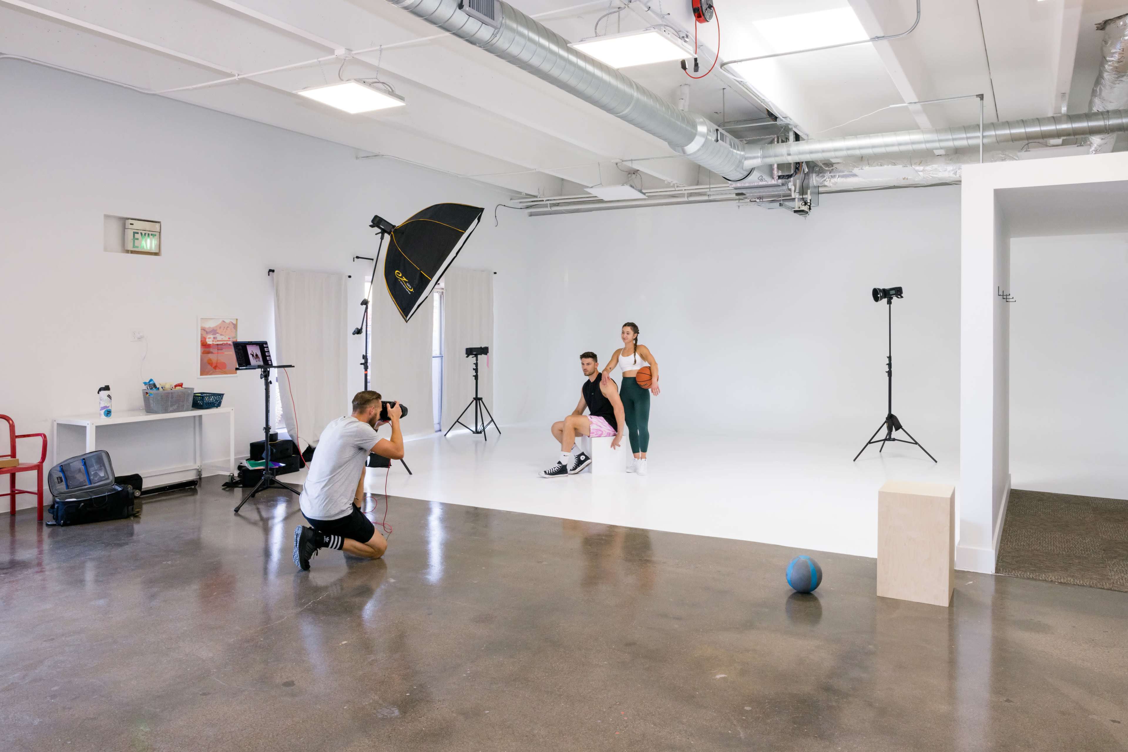 A photographer captures a male and female model in a studio setting with a white backdrop and various lighting equipment.