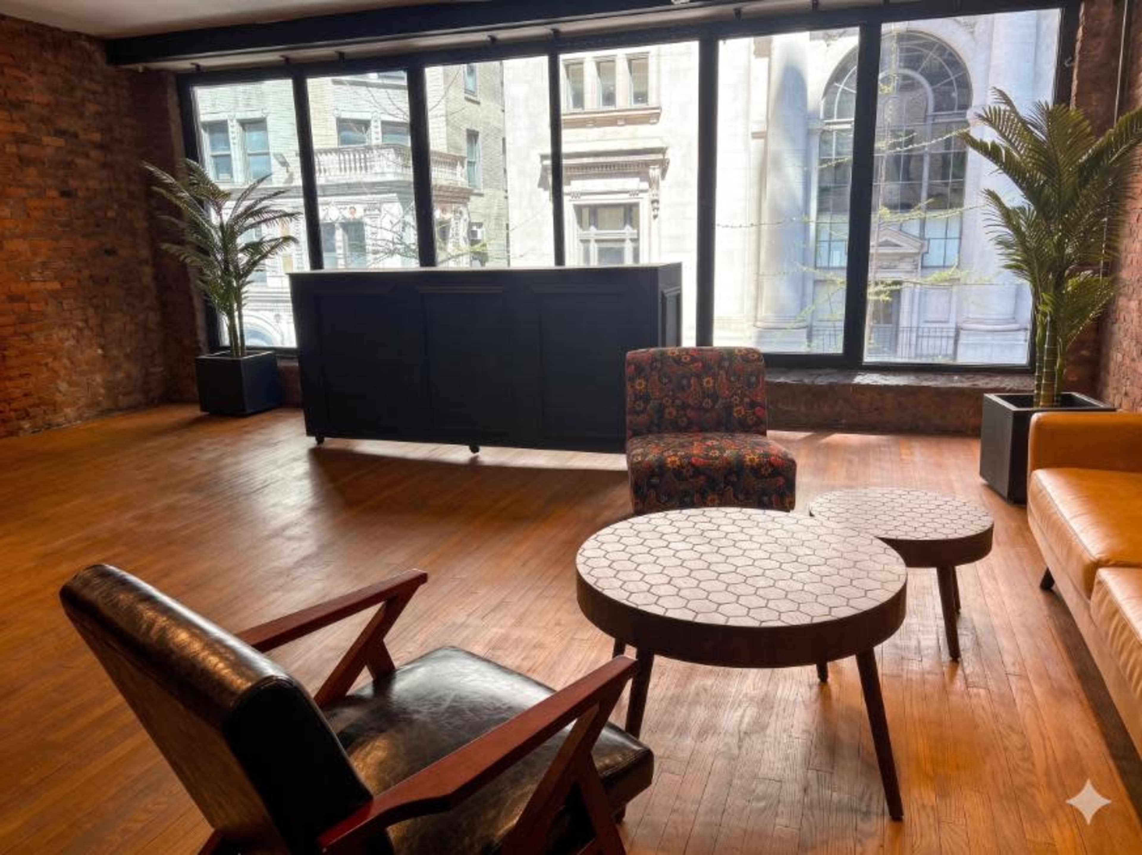 The image shows a modern room with wooden flooring, large windows, and a mix of furniture including two chairs, a coffee table with a hexagonal pattern, and a black partition.