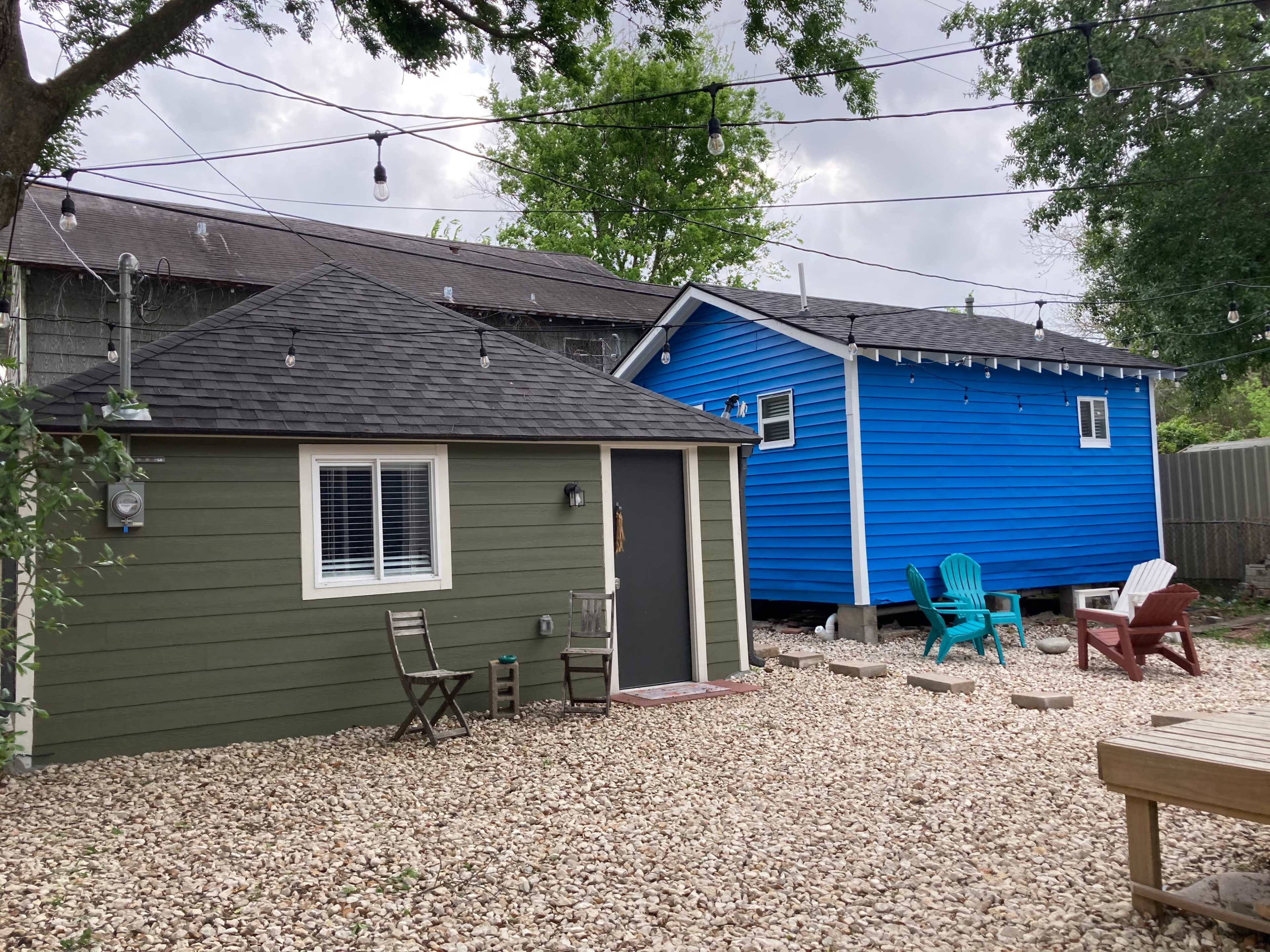 Two houses, one green and one blue, situated next to each other in a gravel yard with outdoor seating and string lights above.