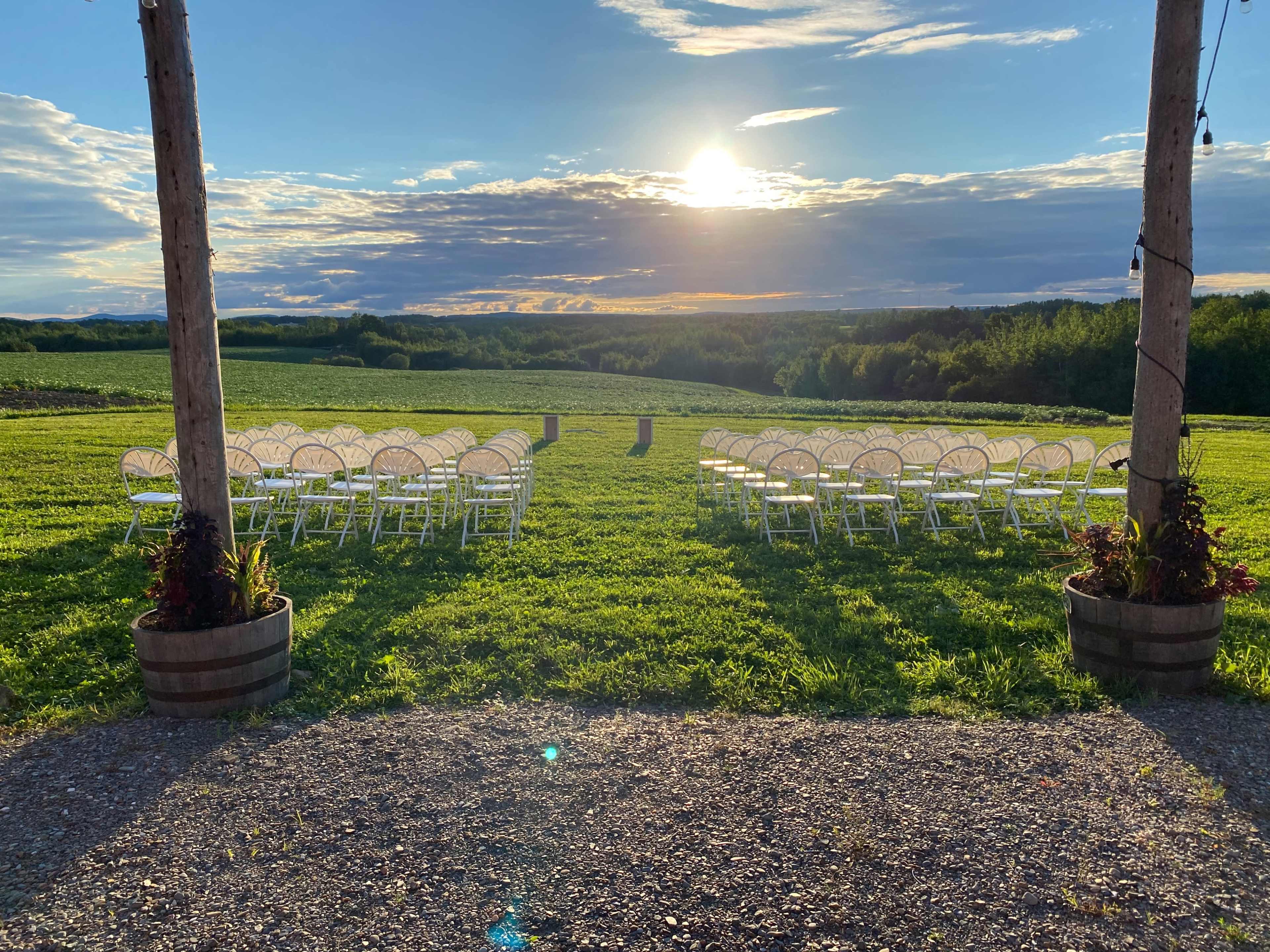A row of white chairs is set up in an open field, facing a sunset over distant hills.