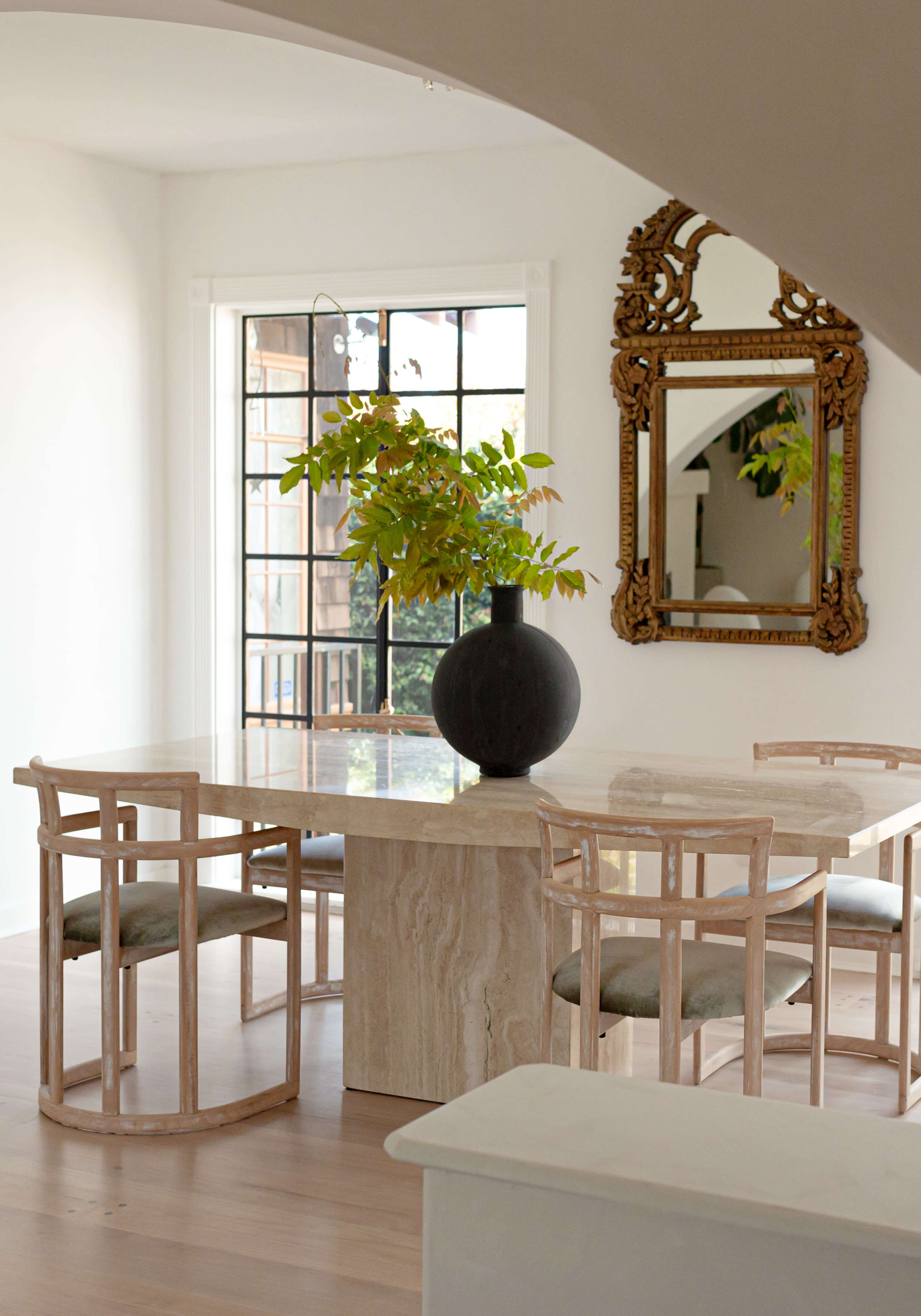 A modern dining area features a stone table surrounded by wooden chairs and a large vase with greenery, complemented by a mirror on the wall.