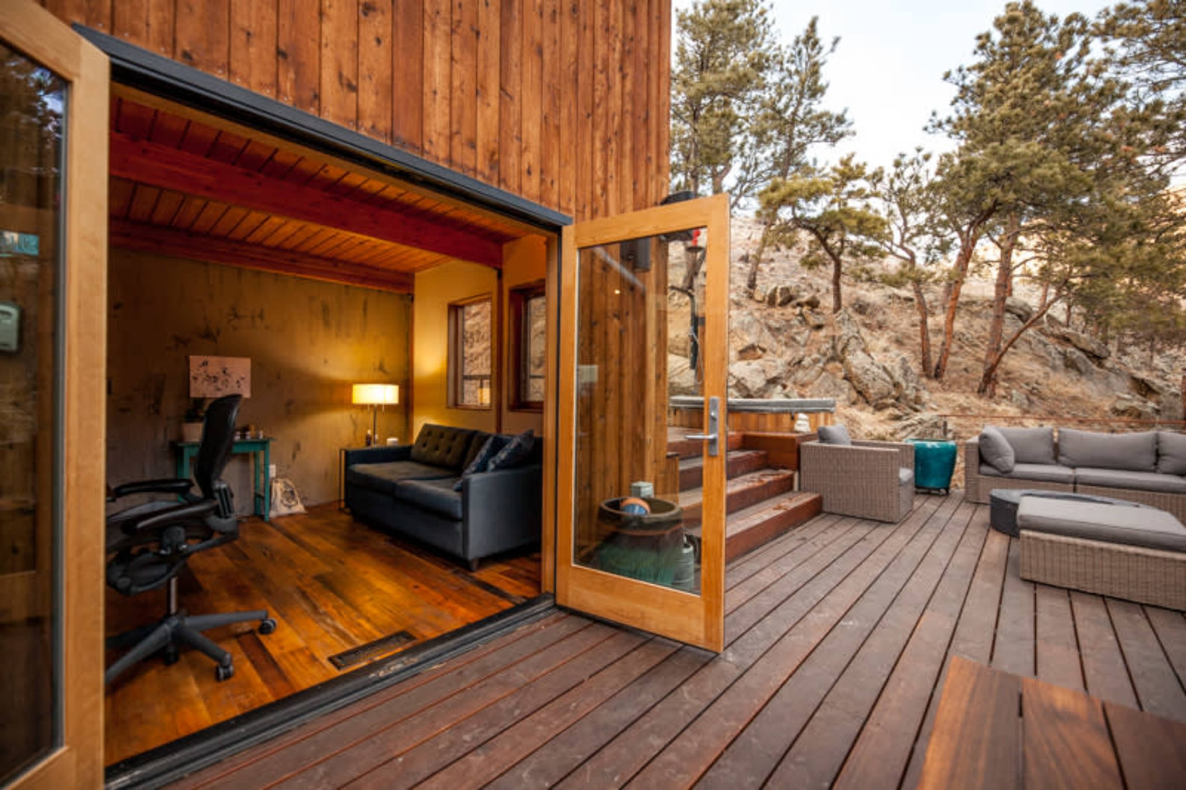 A wooden deck leads to an open door, revealing a cozy interior with a sofa and desk, surrounded by trees and rocky terrain.
