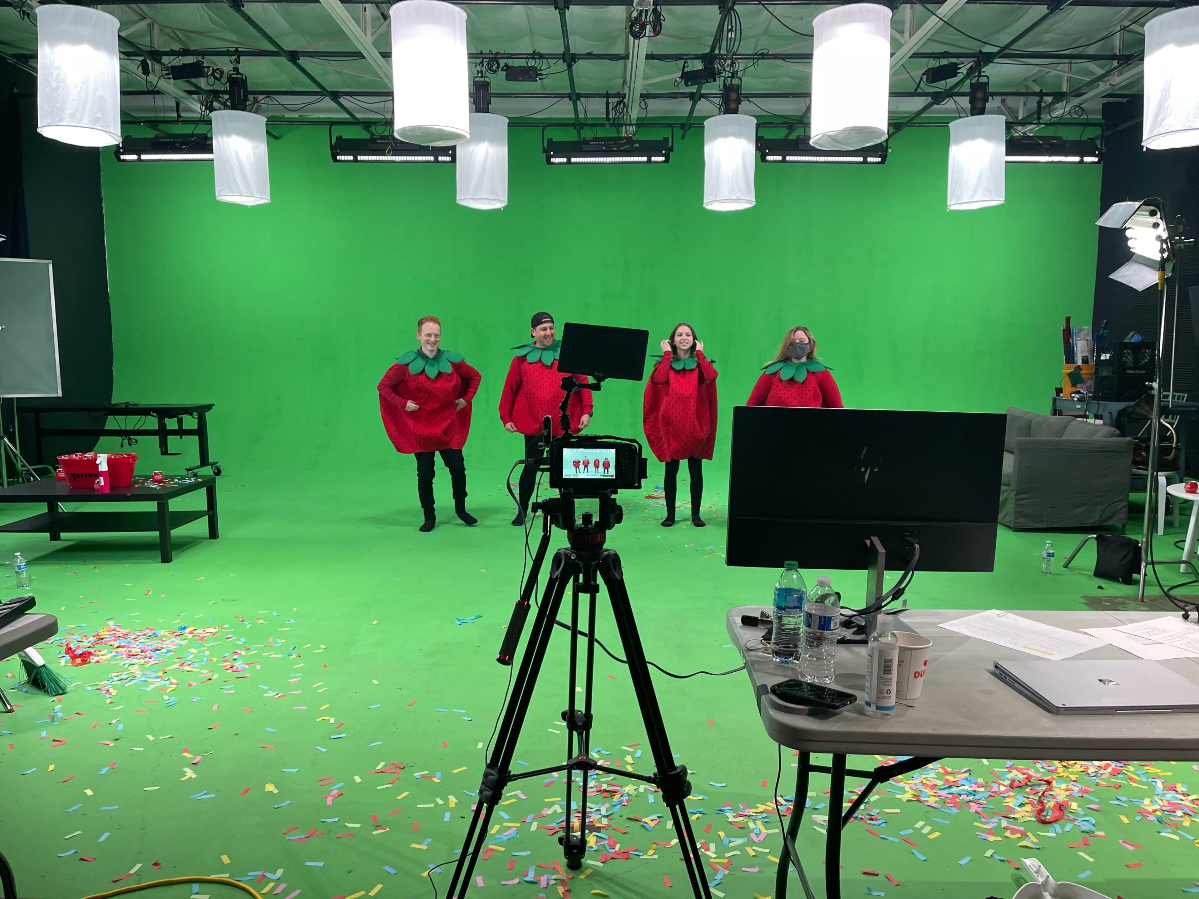 A group of four individuals dressed in red costumes stands in front of a green screen, with a camera setup and colorful confetti scattered on the floor.