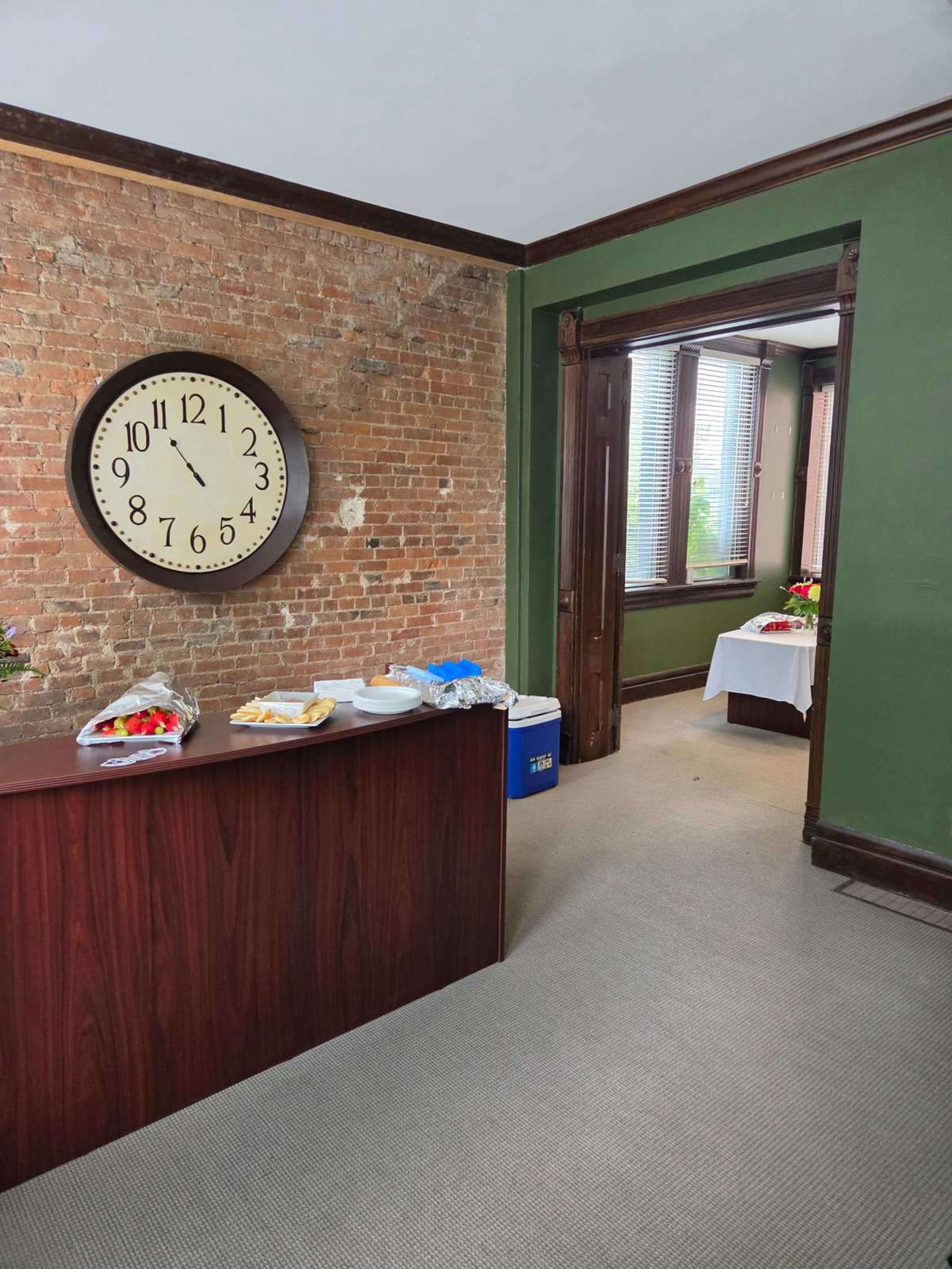 The image shows a room with a exposed brick wall, a large wall clock, and a wooden reception desk beside a doorway leading to another area with tables.