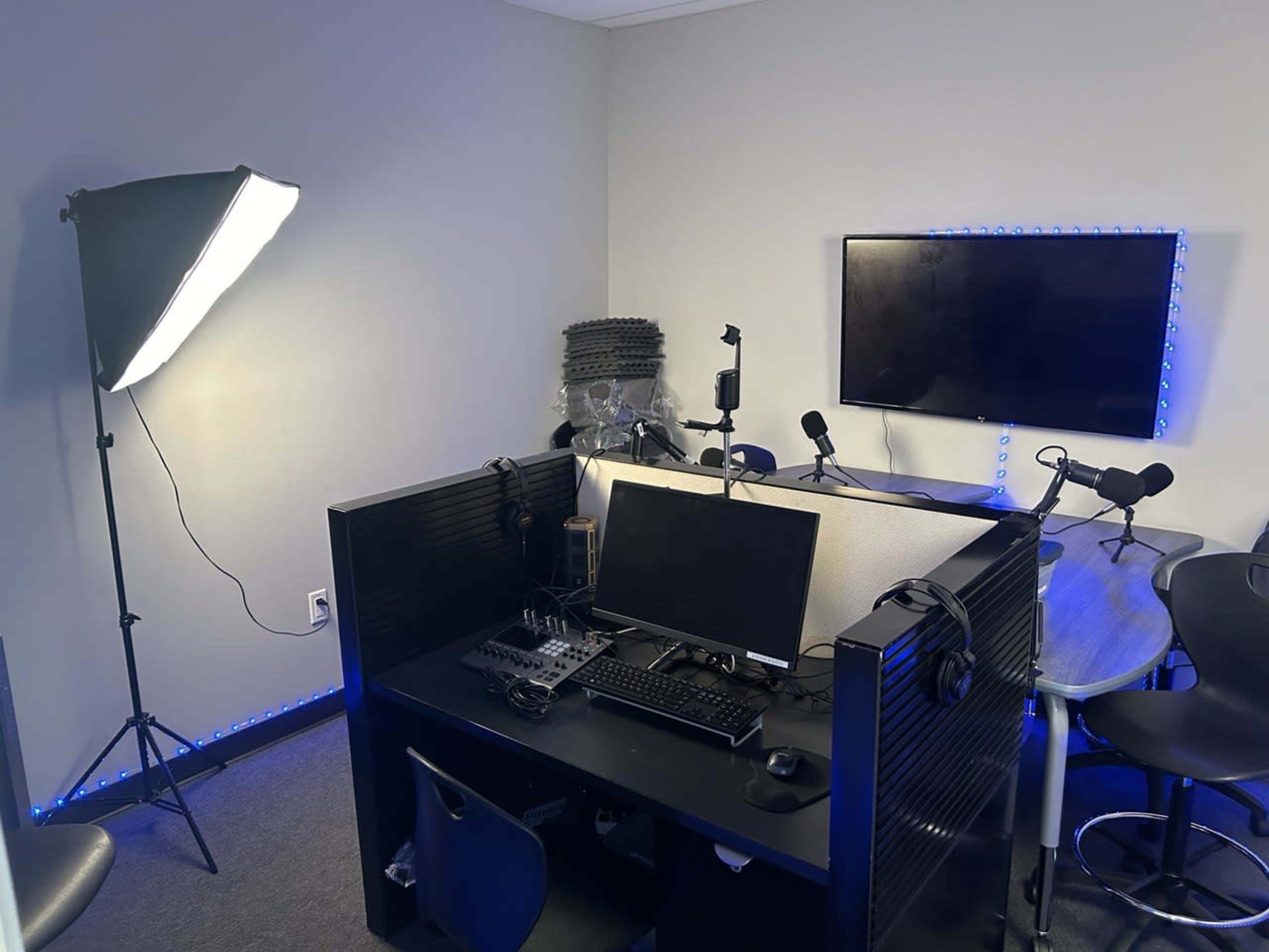 A recording studio setup features a desk with a computer, microphones, and a monitor, accompanied by lighting equipment in a corner of the room.